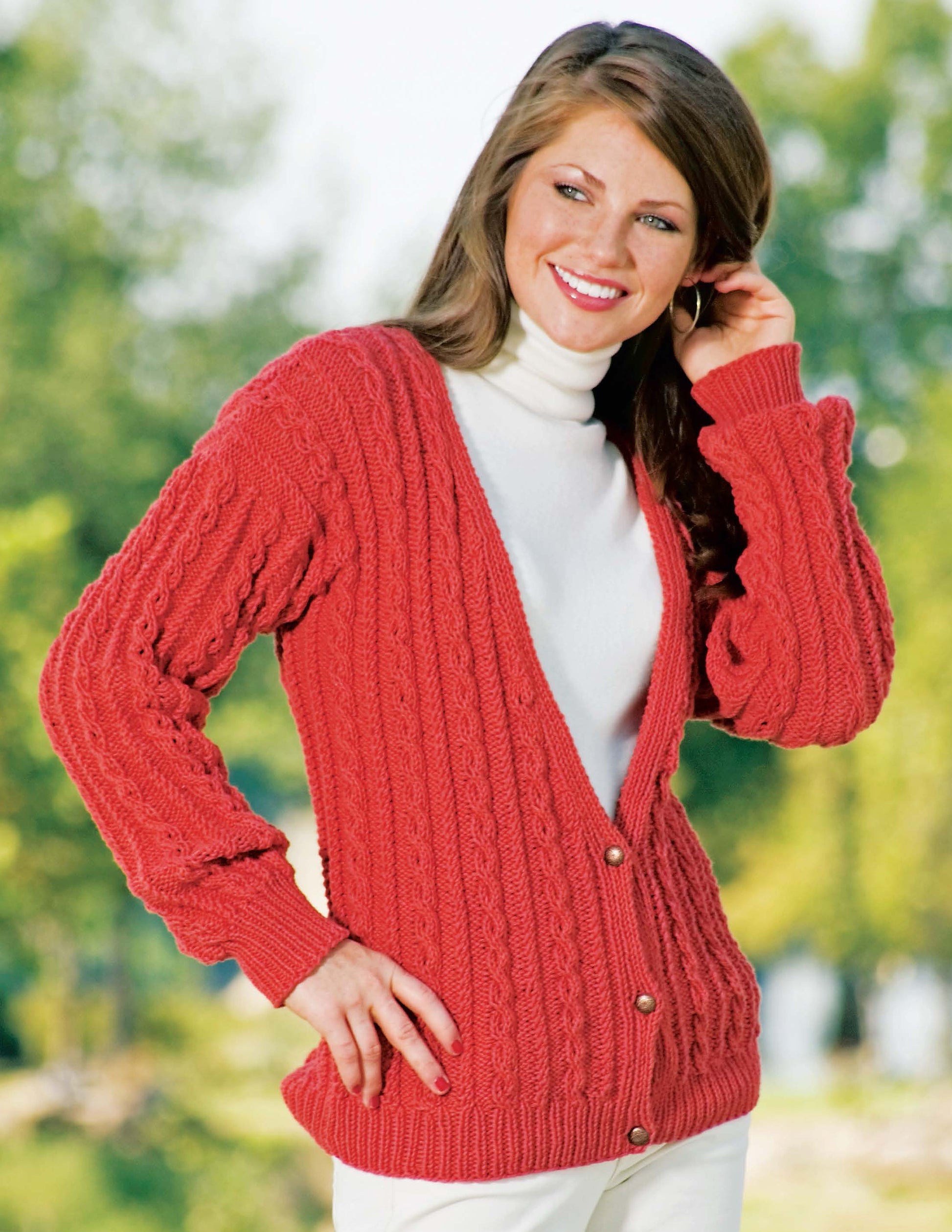 A woman outdoors smiles, touching her hair while wearing a white turtleneck and a red cable-knit cardigan made with Leisure Arts' "I Can't Believe I'm Knitting Cables," paired with white pants against blurred greenery.