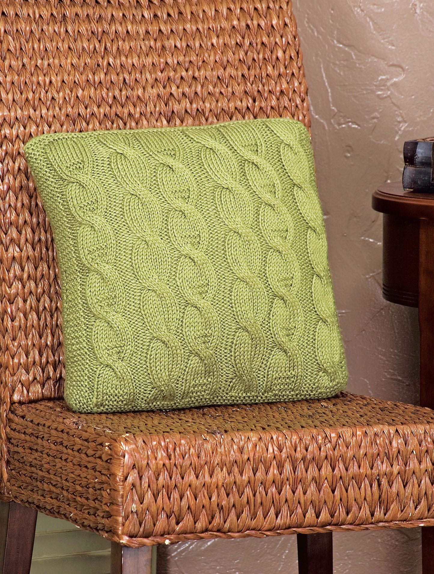 The “I Can’t Believe I’m Knitting Cables” pillow by Leisure Arts, in green with intricate cable patterns, sits on a woven rattan chair beside a wooden table against a textured beige wall.