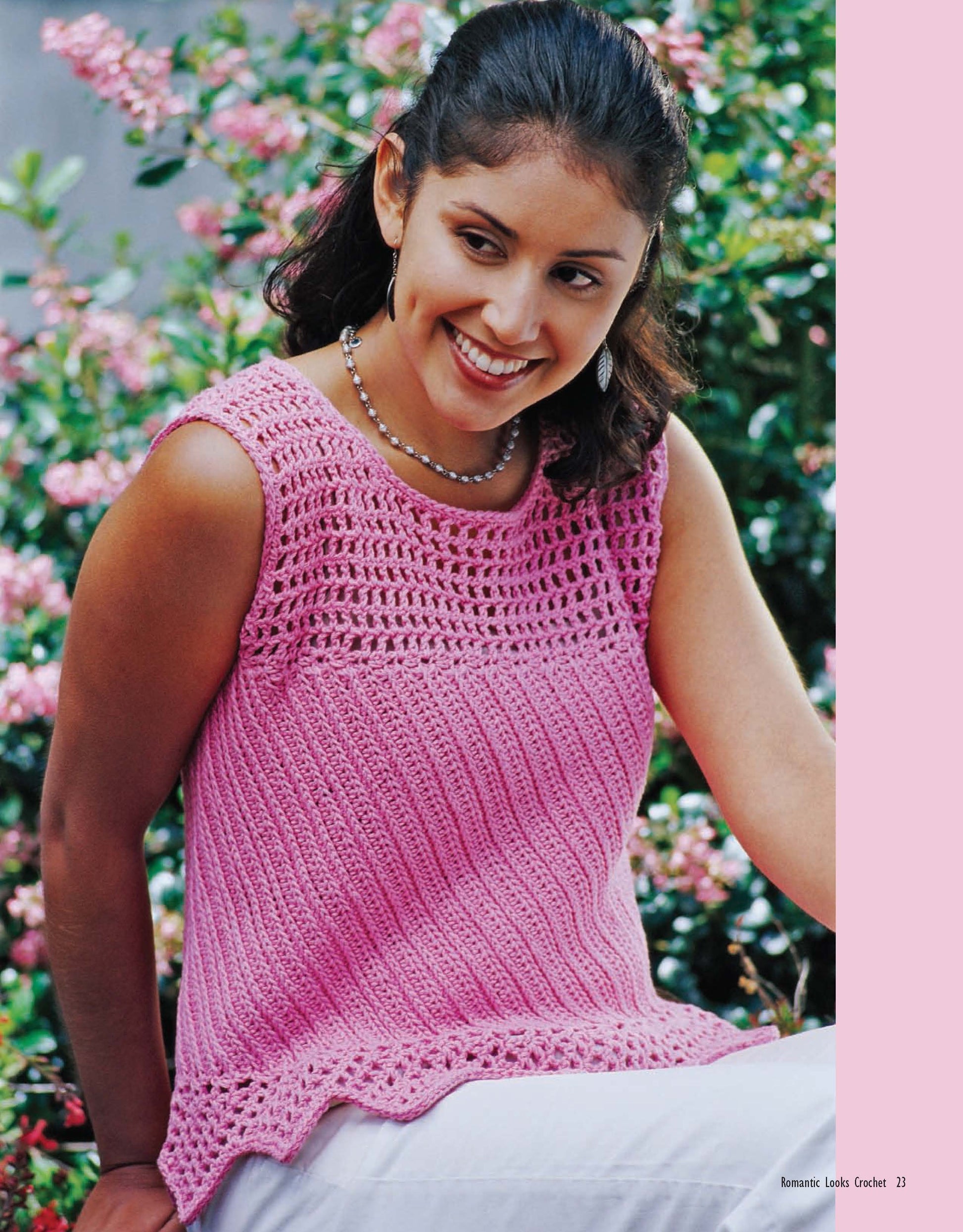 A woman smiles outdoors in front of pink flowers, wearing a sleeveless pink top with a lacy design from Leisure Arts’ Romantic Looks Crochet collection, paired with a beaded necklace.