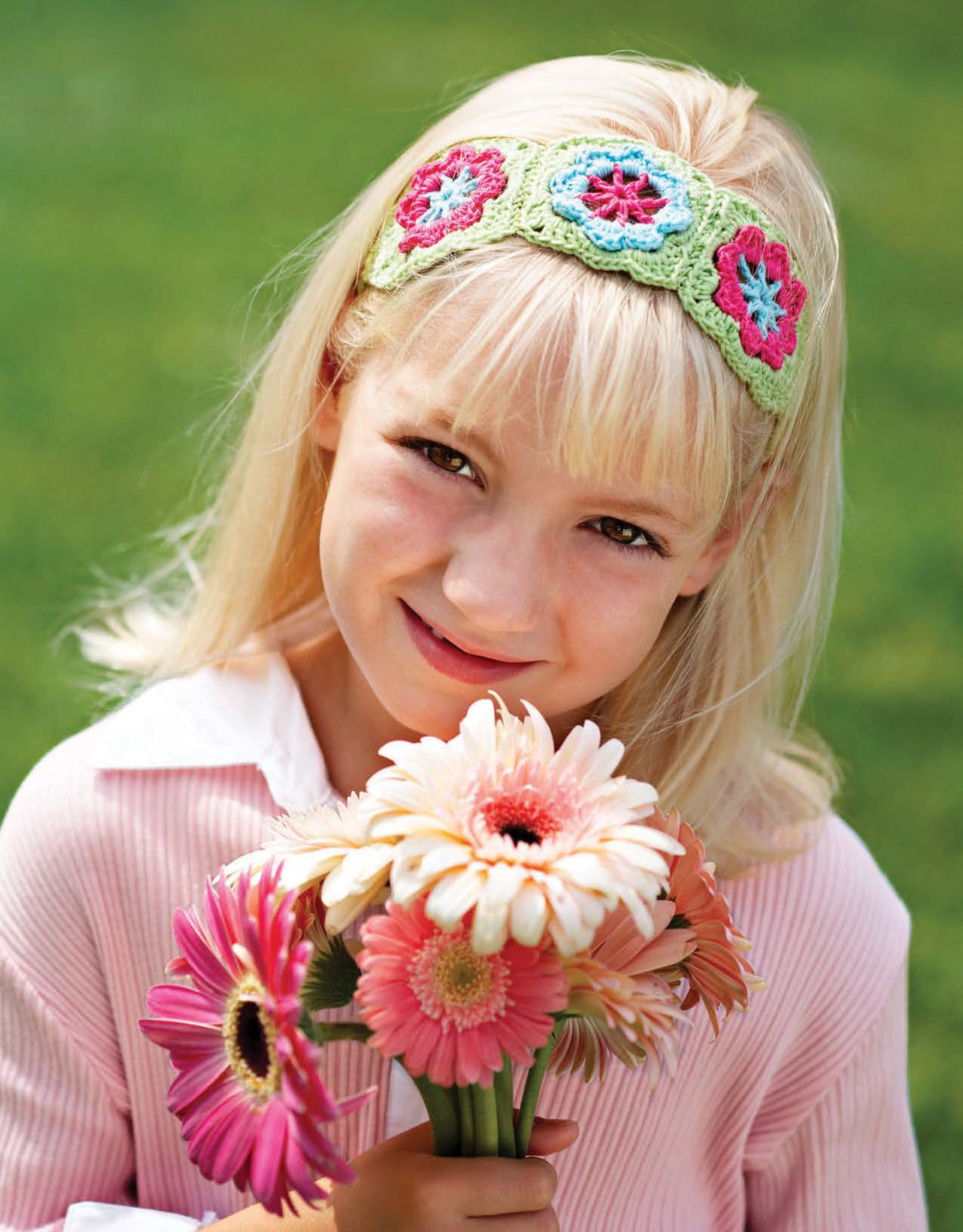 A young blonde girl wearing a crocheted floral headband from Leisure Arts’ Crochet on the Go! smiles, holding a bouquet of colorful gerbera daisies—perfect inspiration for playful beginner crafts with a blurred green grass background.