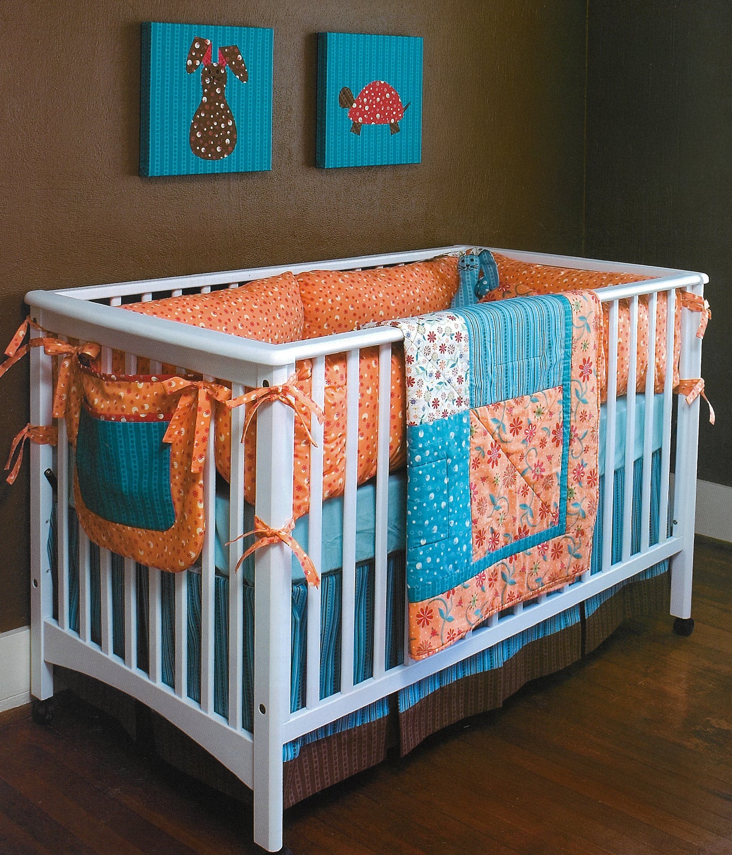 The Leisure Arts iQuilt: No Holds Barred Designers' Mix features a white crib with orange and blue bedding plus a matching quilt, perfect for fat-quarter projects. Above it, blue bunny and turtle wall art accent the wood floors and brown walls.