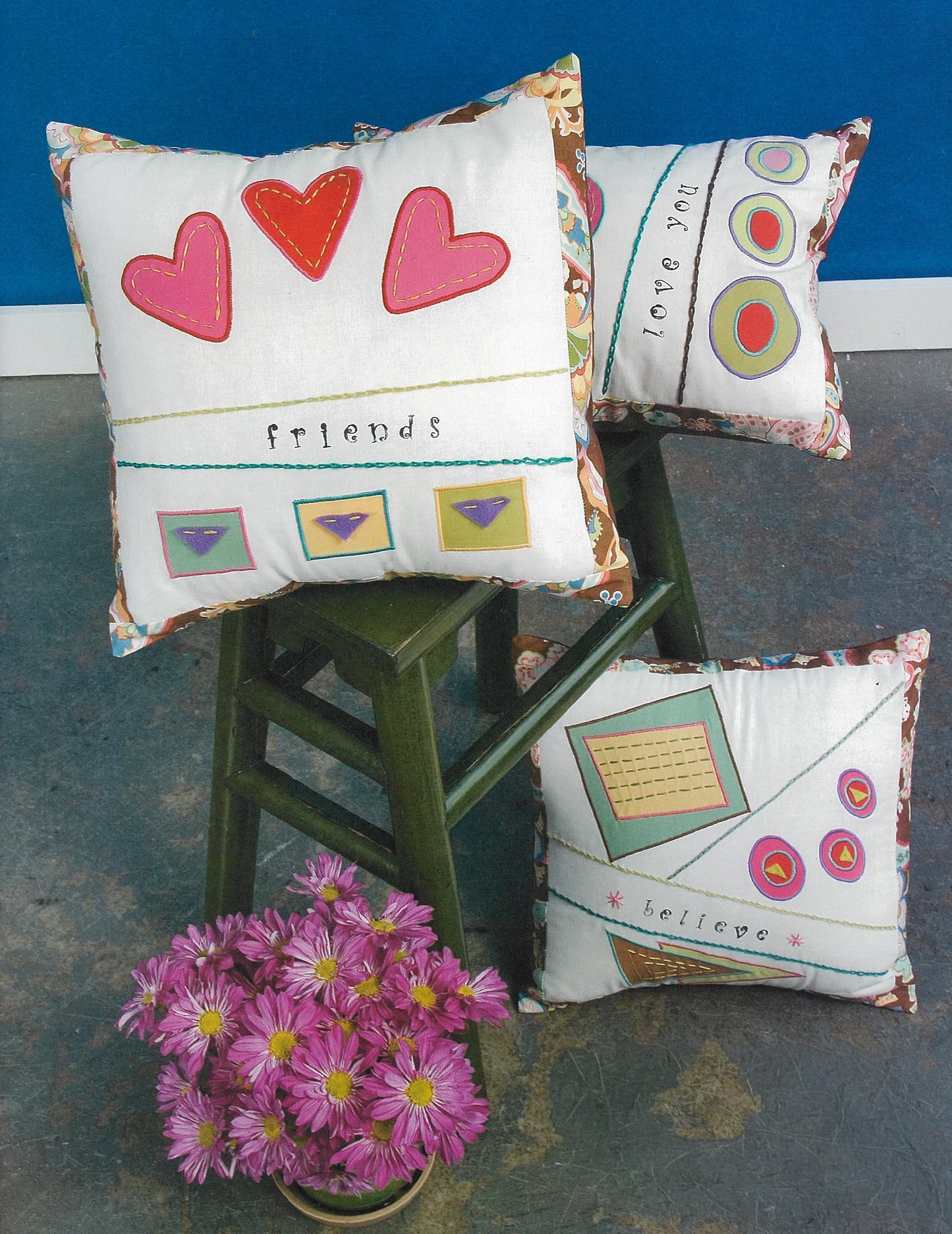 Leisure Arts' iQuilt: No Holds Barred Designers' Mix features three vibrant decorative pillows with geometric and heart patterns, shown on a green chair with a vase of purple flowers against a blue backdrop—ideal for fat-quarter quilt projects.