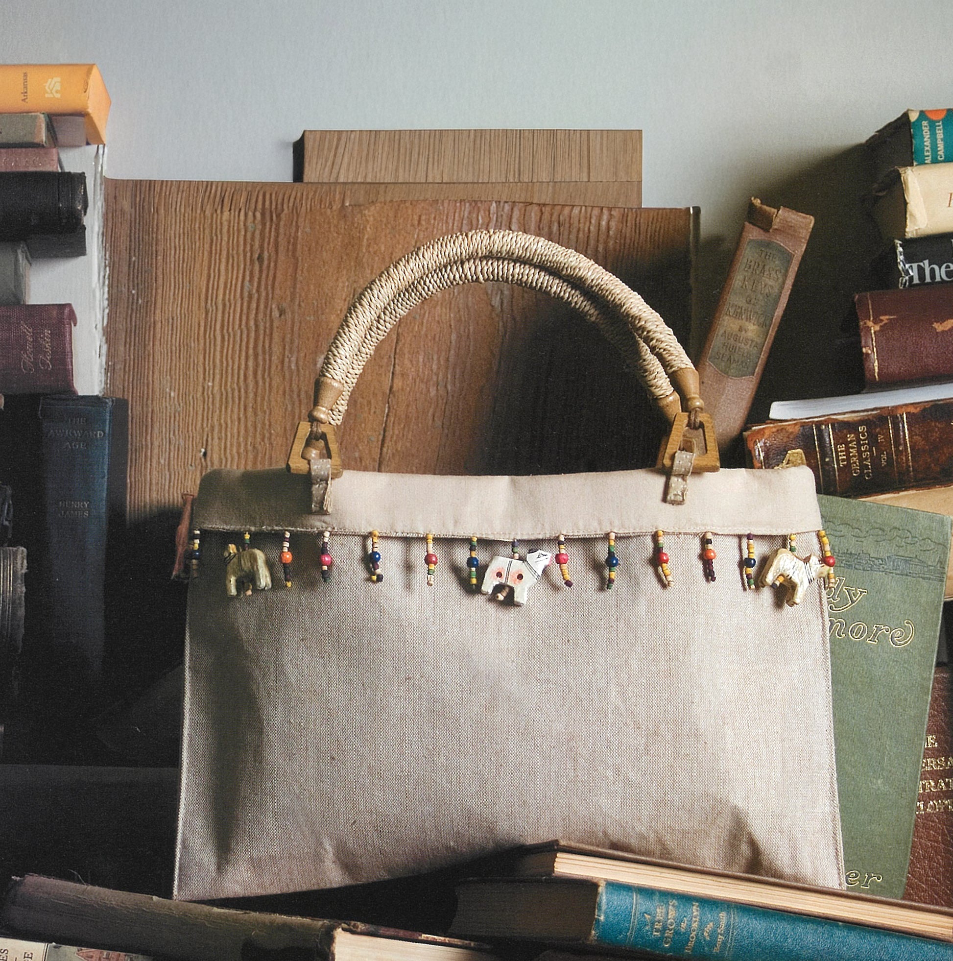 The iQuilt: No Holds Barred Designers' Mix by Leisure Arts sits among vintage books—a beige handbag with a woven handle and colorful charms, perfect for toting instructions or fabric for your next quilting project.