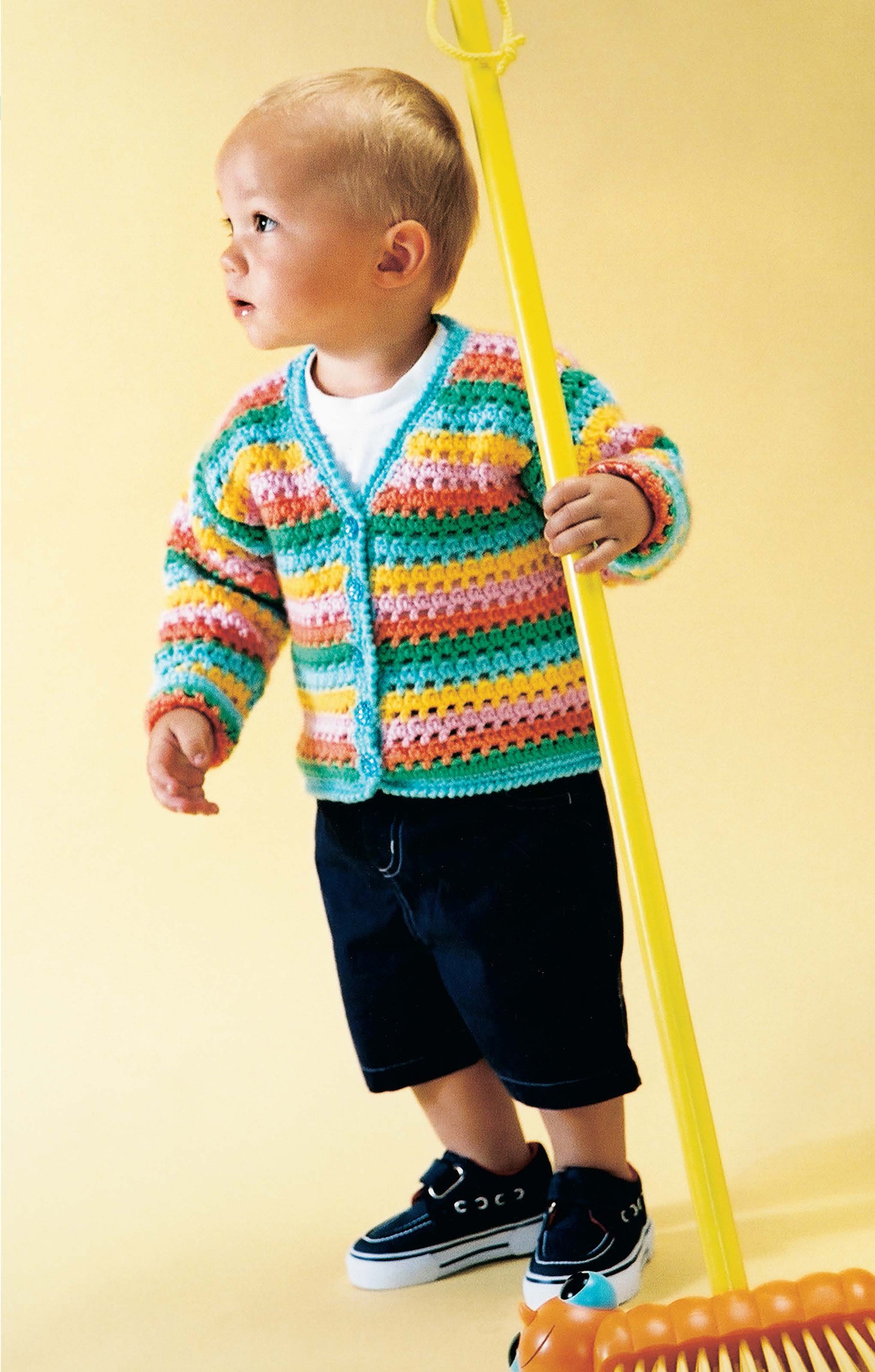 A toddler in a colorful striped knitted cardigan and black shorts stands on a yellow background, holding a large yellow toy rake—a playful moment inspired by Leisure Arts' beginner-friendly guide, 1-2-3 Crochet.