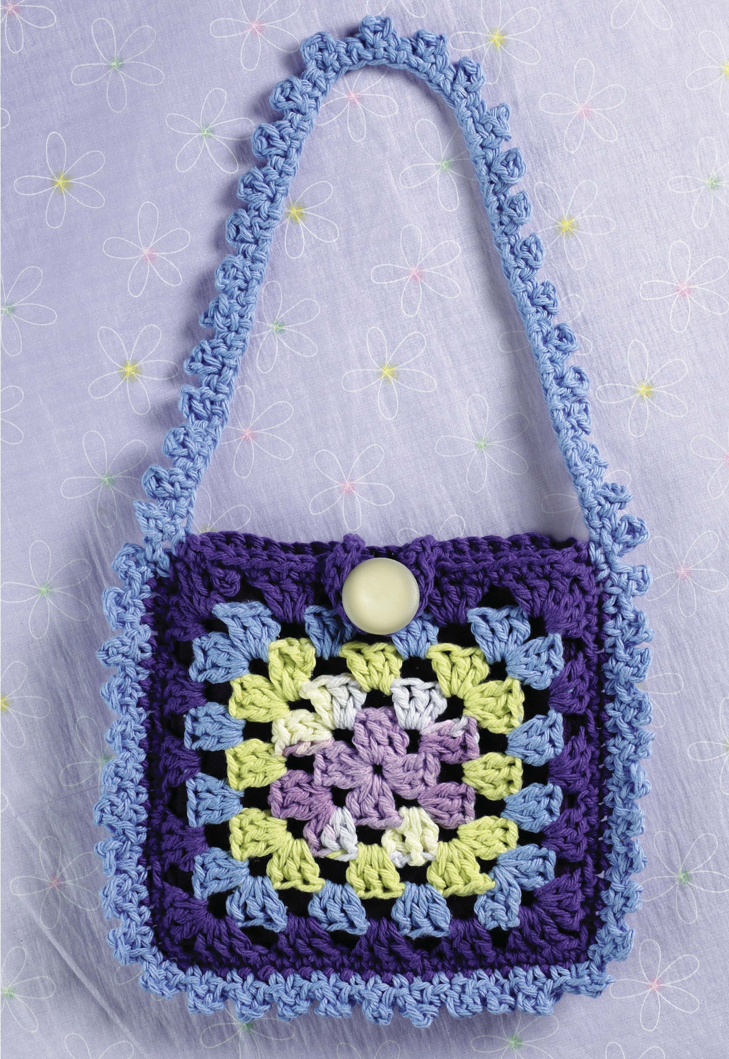 A small crocheted granny square purse in purple, blue, green, lavender, and white with a blue scalloped strap and cream button—made using 1-2-3 Crochet from Leisure Arts—displayed on light purple floral fabric.