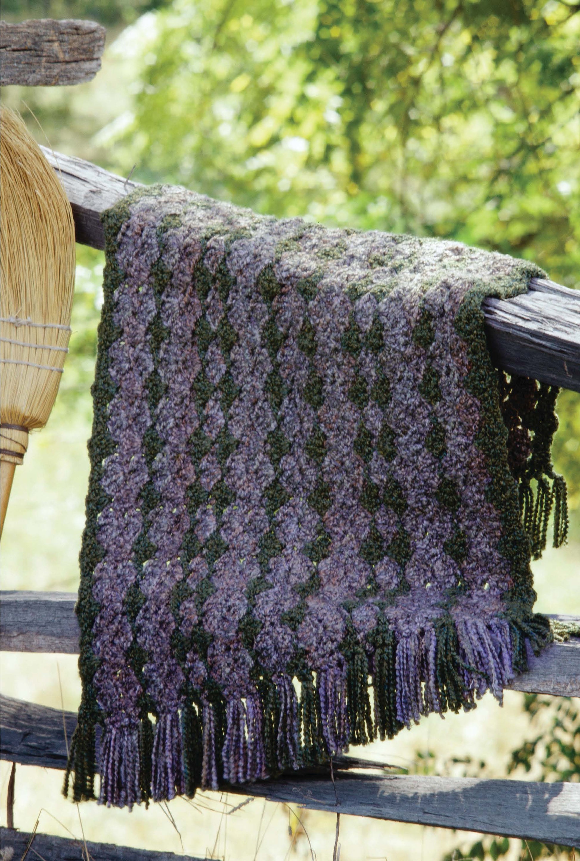A textured purple and green crocheted blanket from Leisure Arts' 1-2-3 Crochet hangs with fringe on a rustic wooden fence outdoors, sunlight and greenery blurred behind. A broom leans nearby.