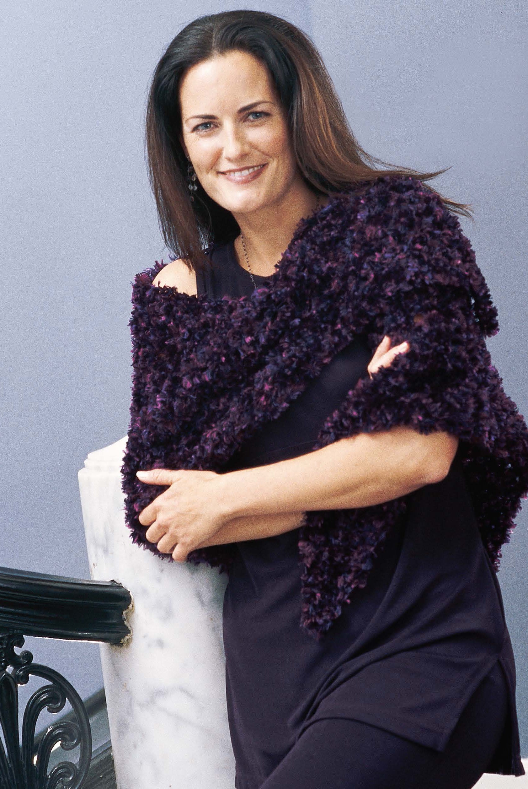 A woman with long brown hair, dressed in a dark purple dress and textured purple shawl, smiles at the camera by a white marble railing—an ideal cover for Leisure Arts' 1-2-3 Crochet beginner's guide. The background is a light purple wall.