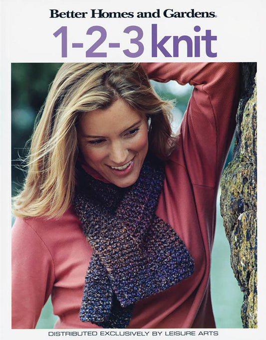 A smiling woman with long blonde hair, in a pink sweater, wears a multicolored knit scarf and leans on a tree. Above her, the text reads: Better Homes and Gardens 1-2-3 Knit by Leisure Arts—a beginner-friendly project.