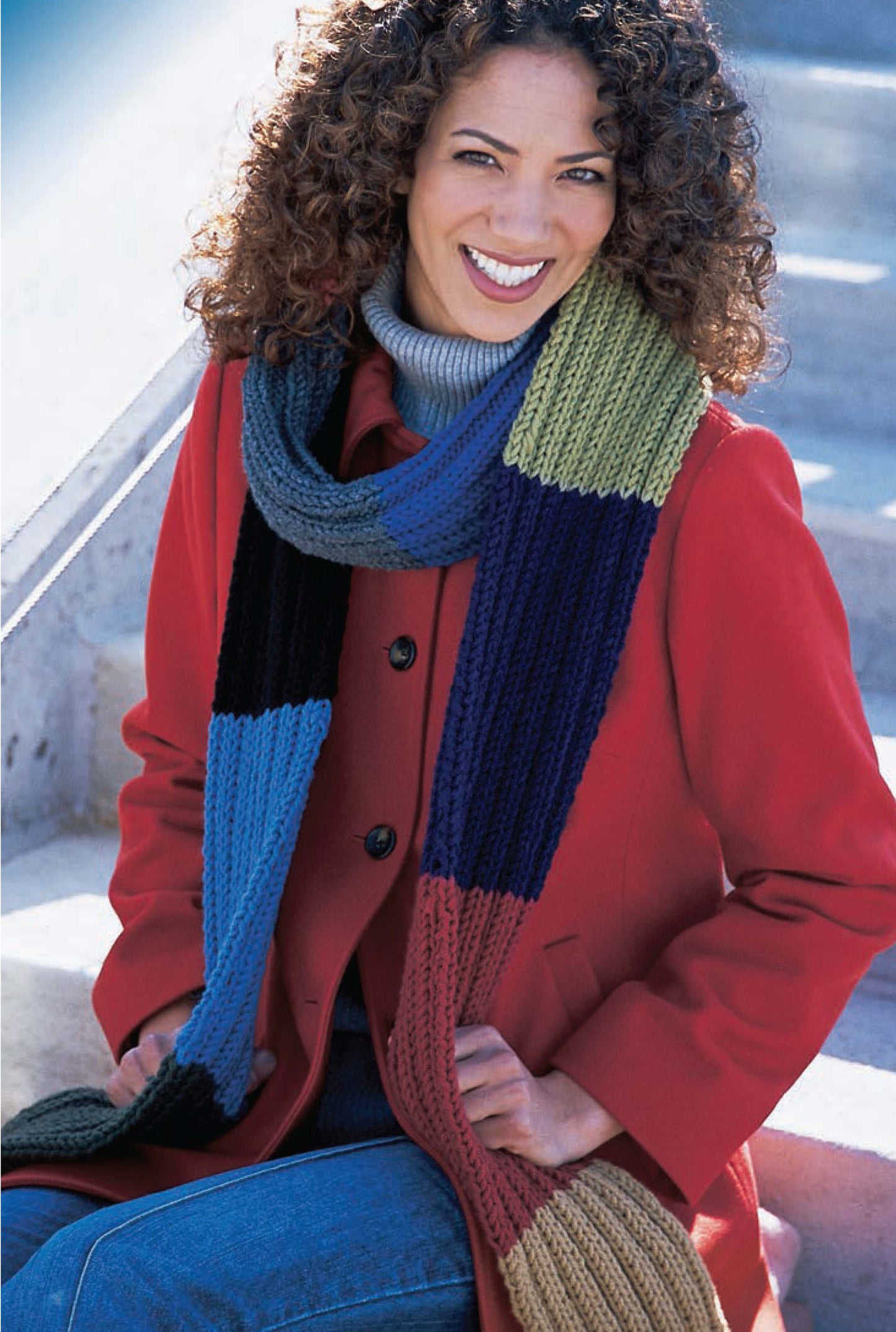 A smiling woman with curly hair wears a red coat, turtleneck, and a long, multicolored scarf from Better Homes and Gardens 1-2-3 Knit by Leisure Arts—a perfect look for beginners enjoying the outdoors.