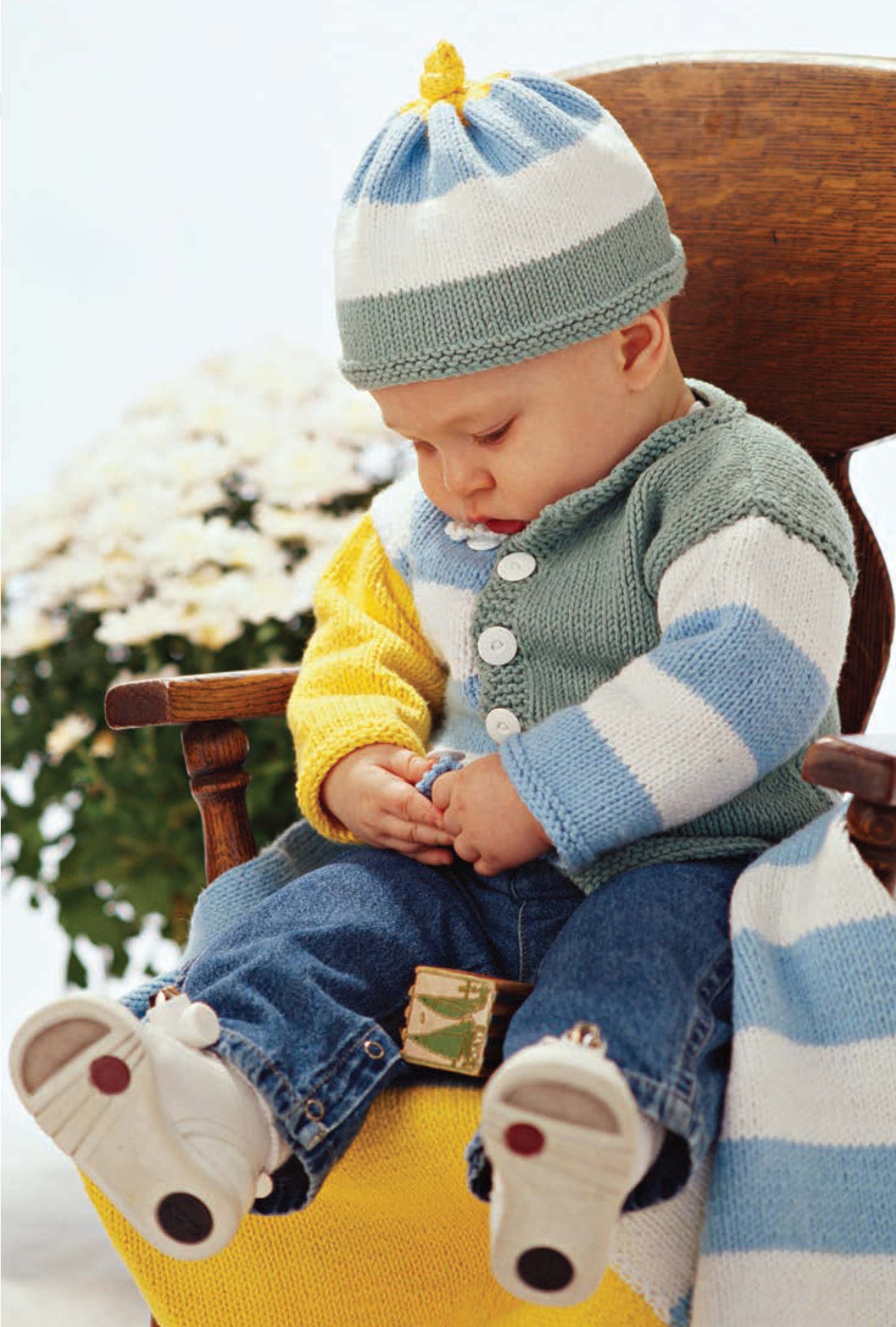 A baby, dressed in a striped knit sweater and matching hat from Better Homes and Gardens 1-2-3 Knit by Leisure Arts, sits on a wooden chair in denim jeans and socks, gazing at their hands with flowers in the background.
