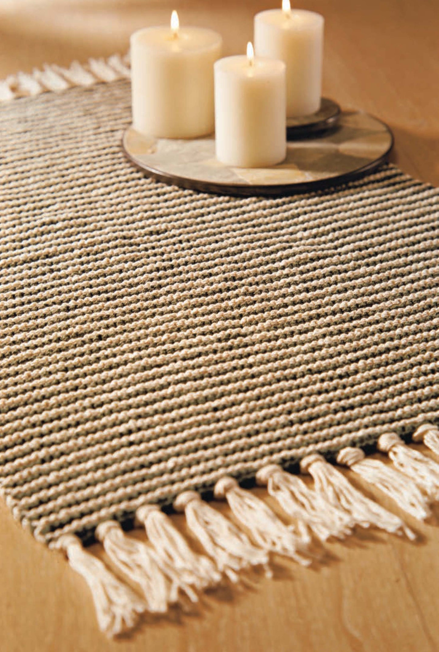 A beige woven table runner from Leisure Arts’ Better Homes and Gardens 1-2-3 Knit collection sits on wood, with three lit pillar candles on a round tray atop the runner for a cozy display.