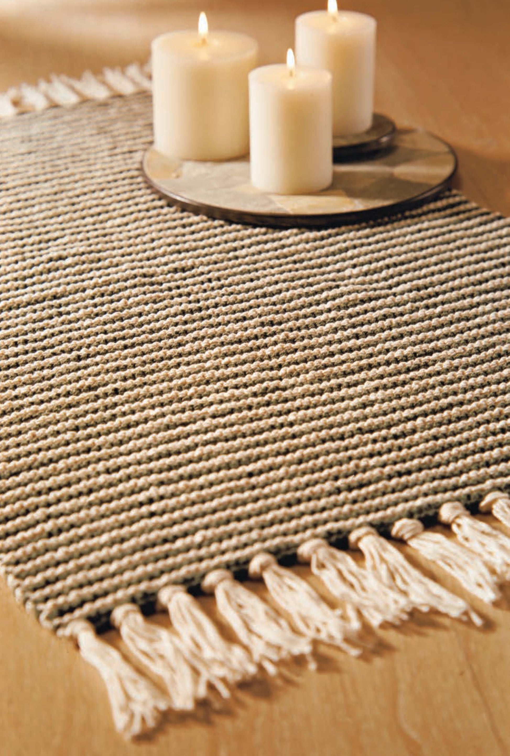 A beige woven table runner from Leisure Arts’ Better Homes and Gardens 1-2-3 Knit collection sits on wood, with three lit pillar candles on a round tray atop the runner for a cozy display.