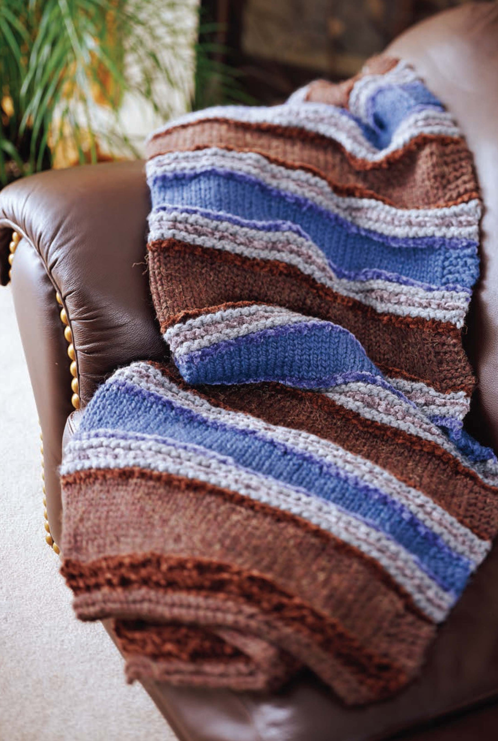 The Better Homes and Gardens 1-2-3 Knit by Leisure Arts, a beginner-friendly blanket with brown, blue, and gray stripes, is draped over a brown leather couch with a leafy plant nearby for a cozy touch.