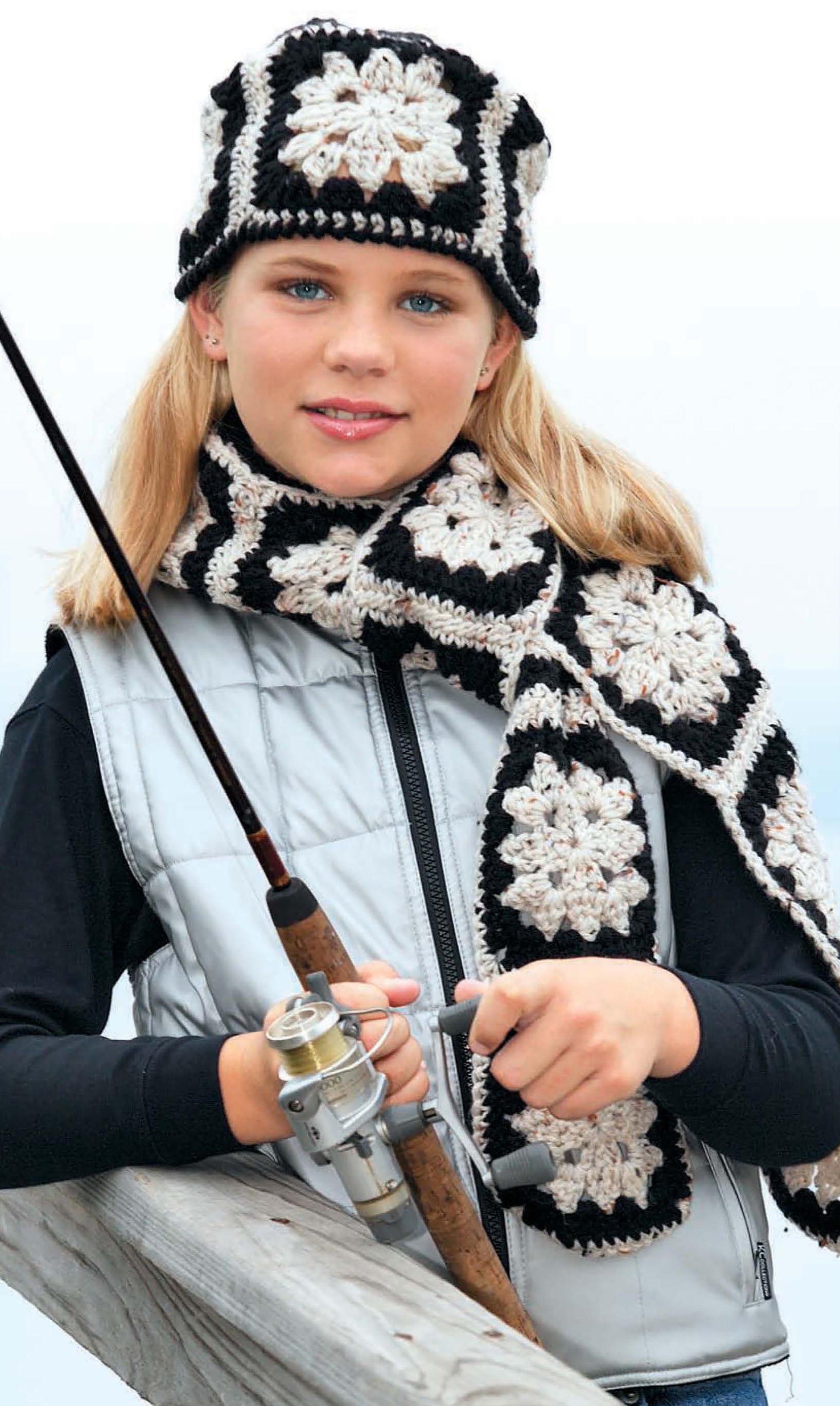 A girl with long blonde hair wears black, white, and grey crocheted chill chasers—a hat and scarf—from the "No Adults Allowed! Digital Download" by Leisure Arts, leaning on a wooden railing in a light grey vest while holding a fishing rod.