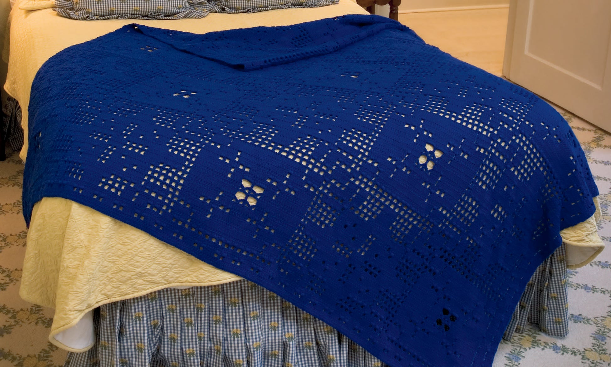 A neatly made bed showcases a blue, patterned crocheted blanket from "Afghans for All Reasons & All Seasons Digital Download" by Leisure Arts, draped over a yellow quilt with a checked bed skirt in a cozy, well-lit bedroom.