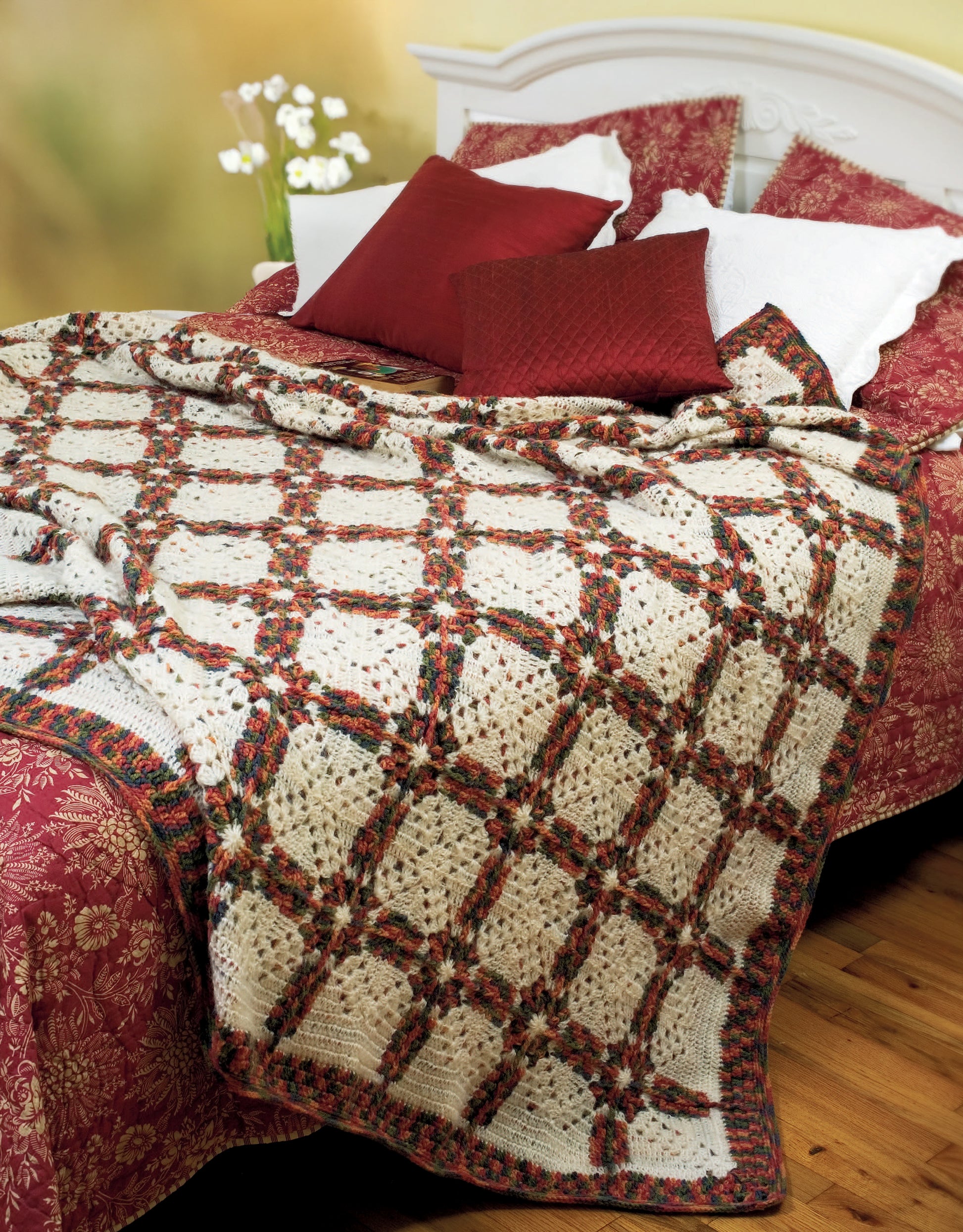 A bed with a white headboard, red and white bedding, and pillows is warmed by a crocheted afghan featuring red, green, and cream lattice patterns made using "Afghans for All Reasons & All Seasons Digital Download" from Leisure Arts.