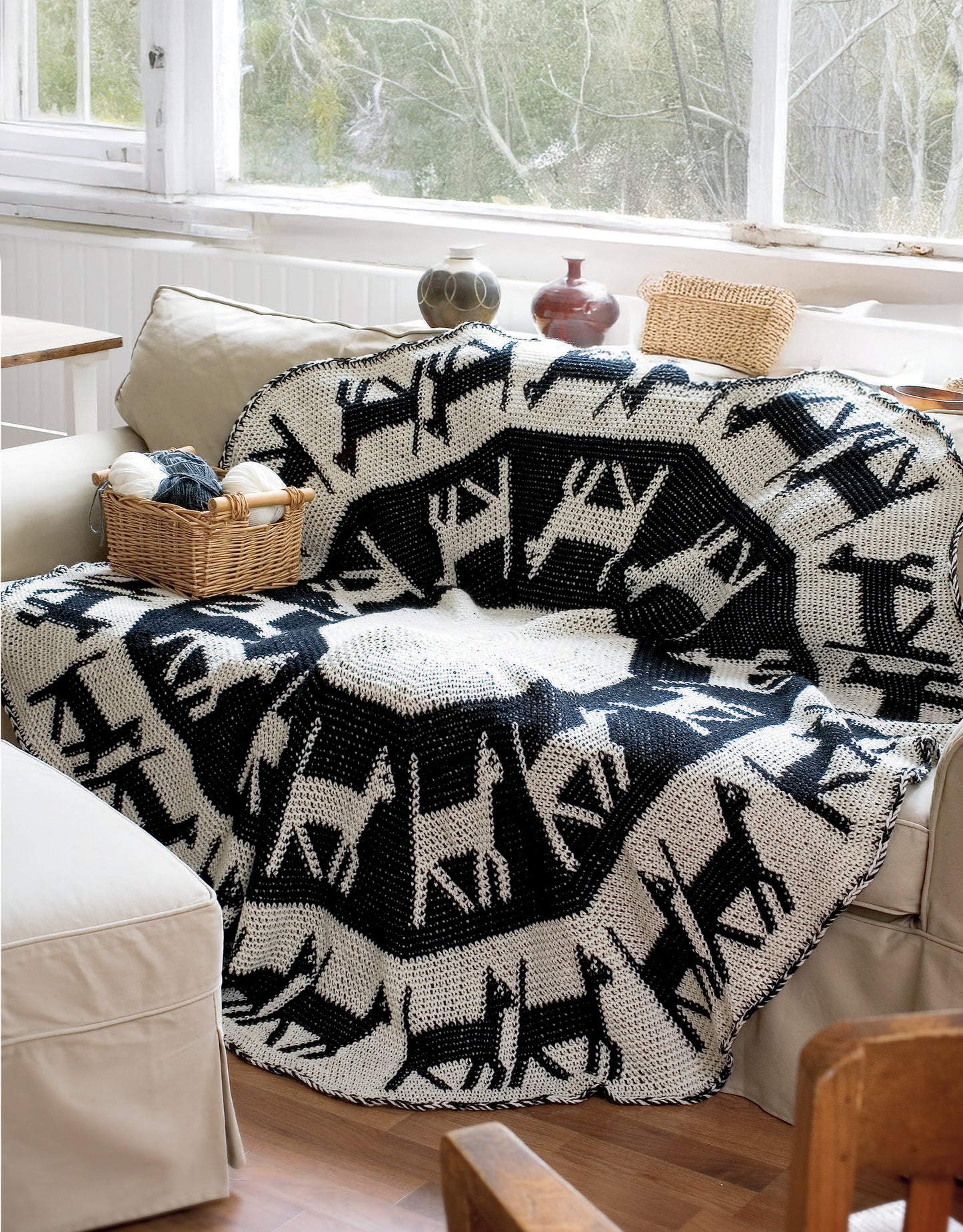 A beige couch displays a black and white crocheted blanket with dog silhouettes from Leisure Arts' "Afghans for All Reasons & All Seasons" digital download, plus yarn supplies on the armrest. Large windows and trees are seen in the background.