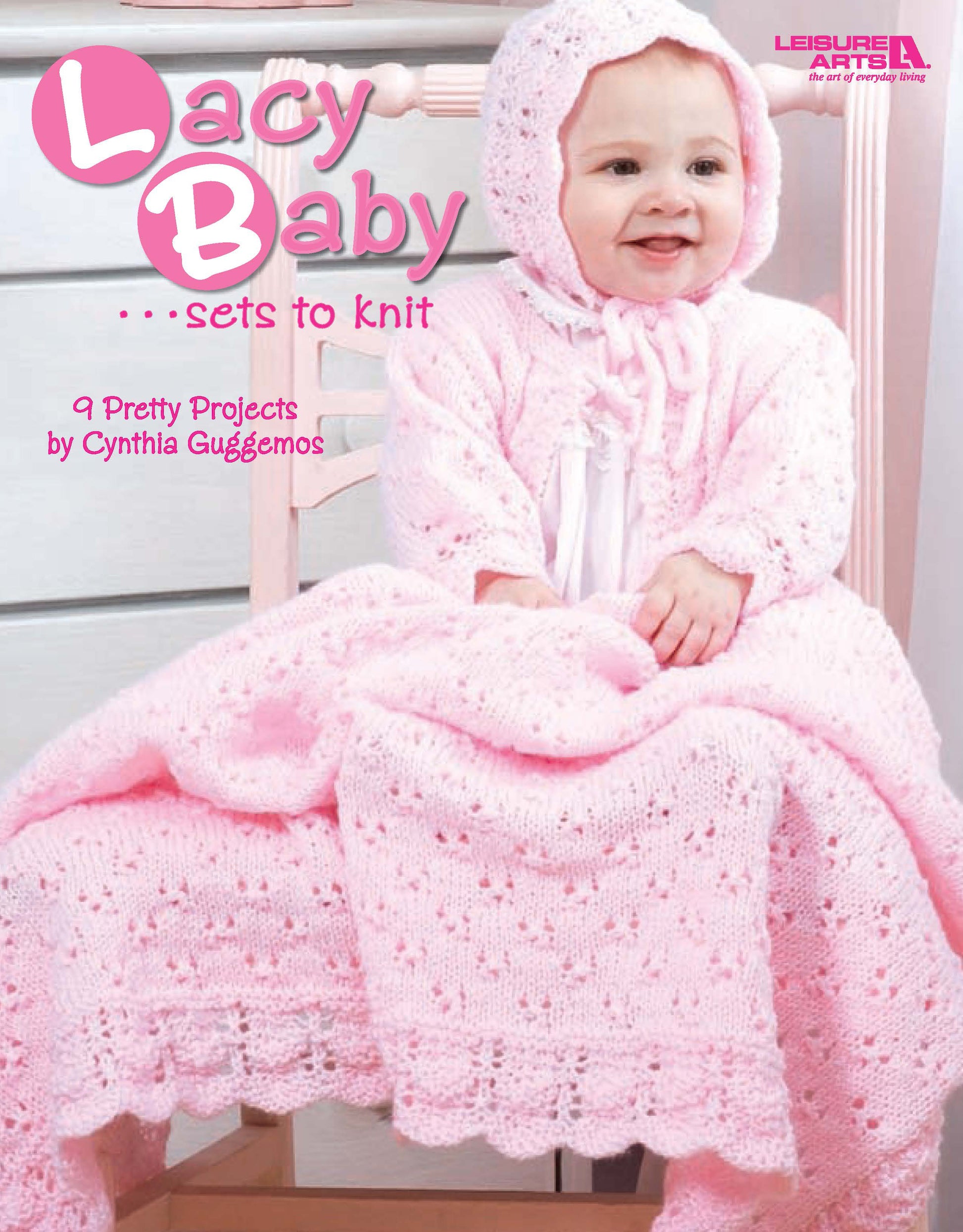 A smiling baby in a pink knitted bonnet and gown sits on a white chair draped with a matching lacy blanket. Text: Lacy Baby Sets to Knit by Leisure Arts – 9 pretty projects for infants designed by Cynthia Guggemos.