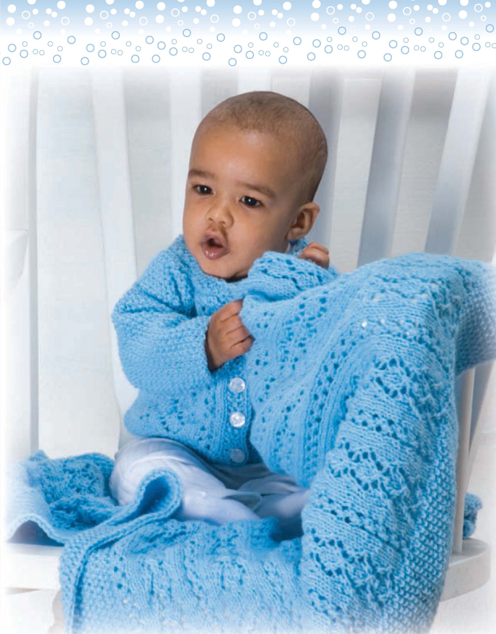 A baby in a light blue sweater sits on a white chair, holding an infant blanket. The snowy white background is perfect for highlighting "Lacy Baby Sets to Knit" by Leisure Arts, ideal for showcasing charming baby knitting patterns.