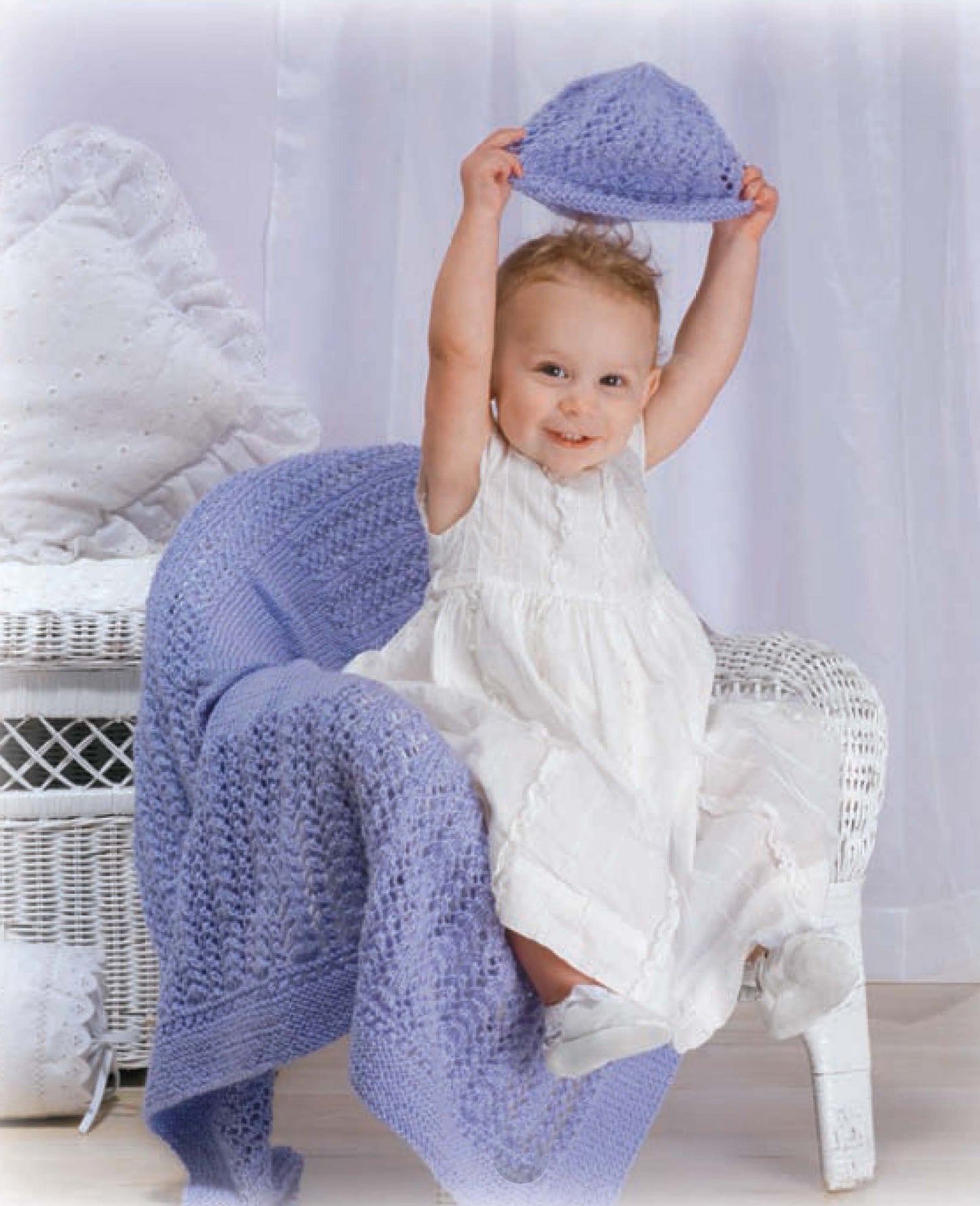 A smiling baby in a white dress sits on a white wicker chair draped with a lavender set from Lacy Baby Sets to Knit by Leisure Arts, holding the matching knitted hat overhead.