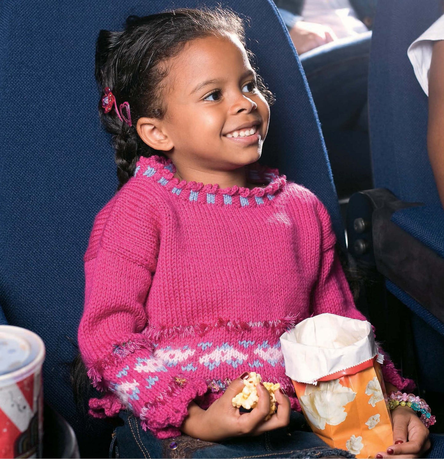 A young girl beams in a theater seat wearing a Leisure Arts Fun Time Sweater, holding popcorn and a colorful bag on her lap, with a drink beside her for the perfect cozy outing.