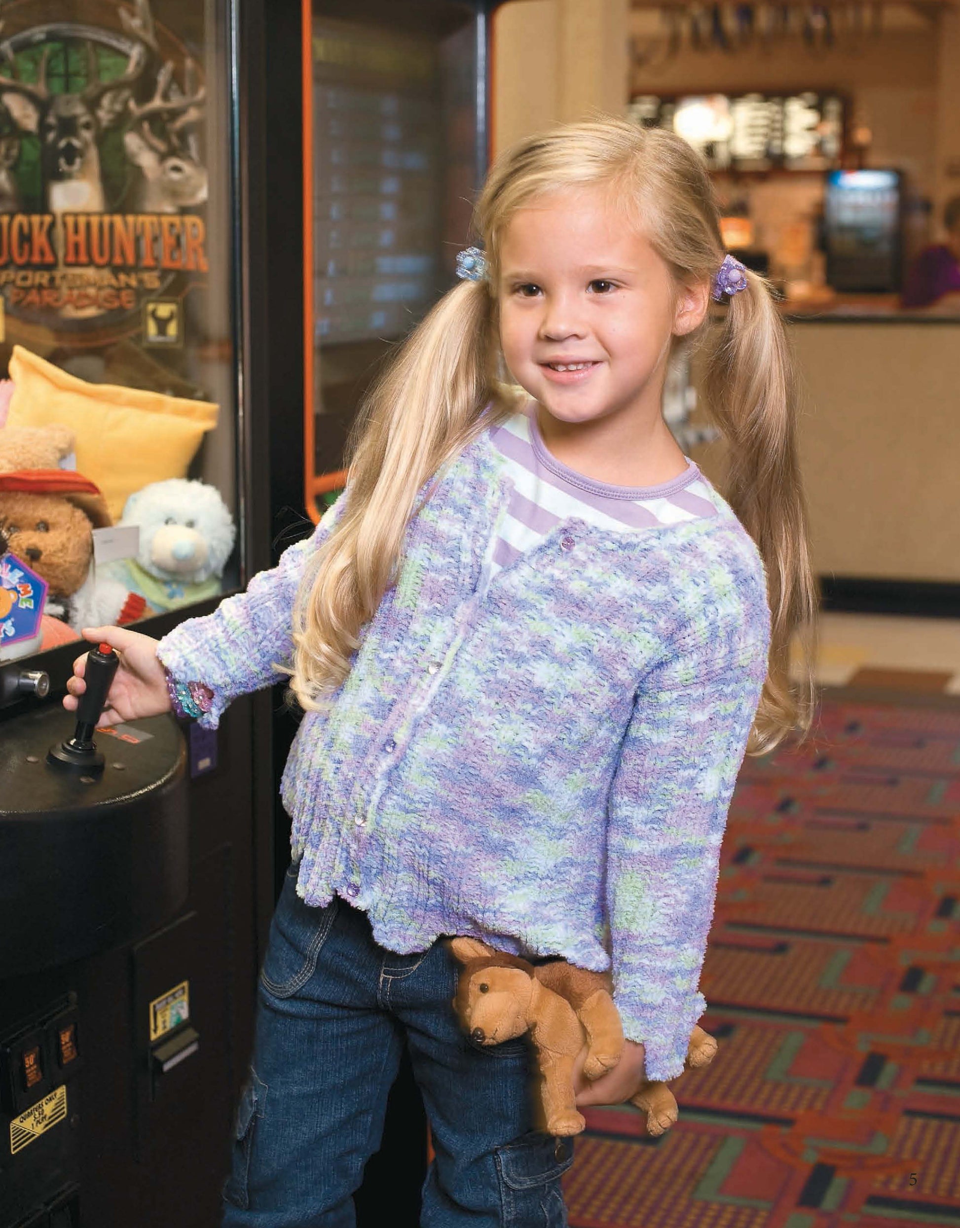 A smiling young girl with blonde pigtails plays a claw machine in an arcade, holding a plush dog. She wears a light purple Fun Time Sweater by Leisure Arts, with more stuffed toys visible inside the machine.