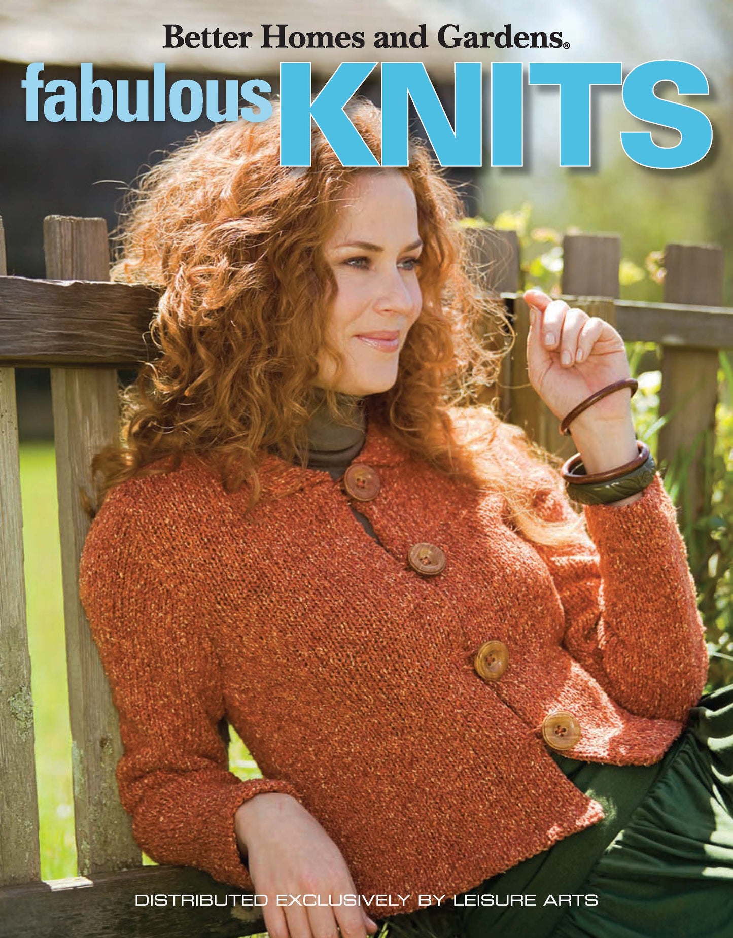 A woman with curly hair, in an orange knit sweater with large buttons, sits outdoors by a wooden fence. Text: Better Homes and Gardens Fabulous Knits by Leisure Arts.