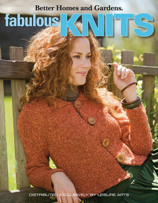 A woman with curly hair, in an orange knit sweater with large buttons, sits outdoors by a wooden fence. Text: Better Homes and Gardens Fabulous Knits by Leisure Arts.