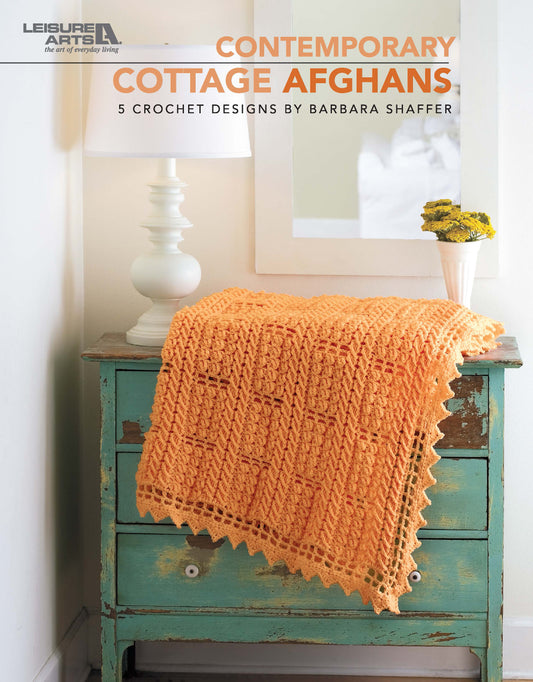 A soft orange worsted weight crocheted afghan is draped over a turquoise nightstand beside a lamp and yellow bouquet, promoting the Leisure Arts “Contemporary Cottage Afghans Digital Download.”.