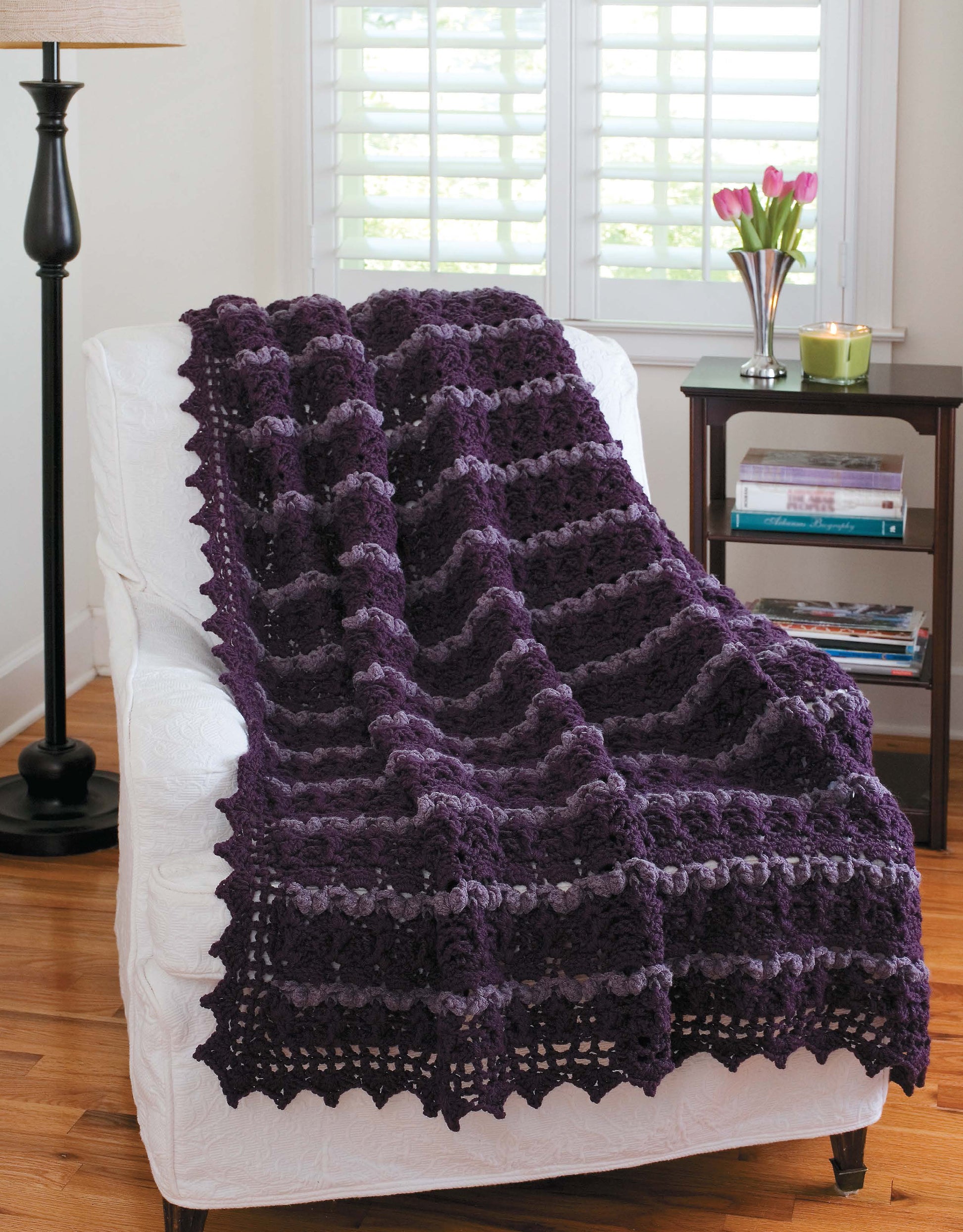The Leisure Arts "Contemporary Cottage Afghans" digital download features a dark purple, worsted weight crocheted blanket with an intermediate pattern, shown draped on a white chair near a black table with books, candle, tulips, and bright window light.