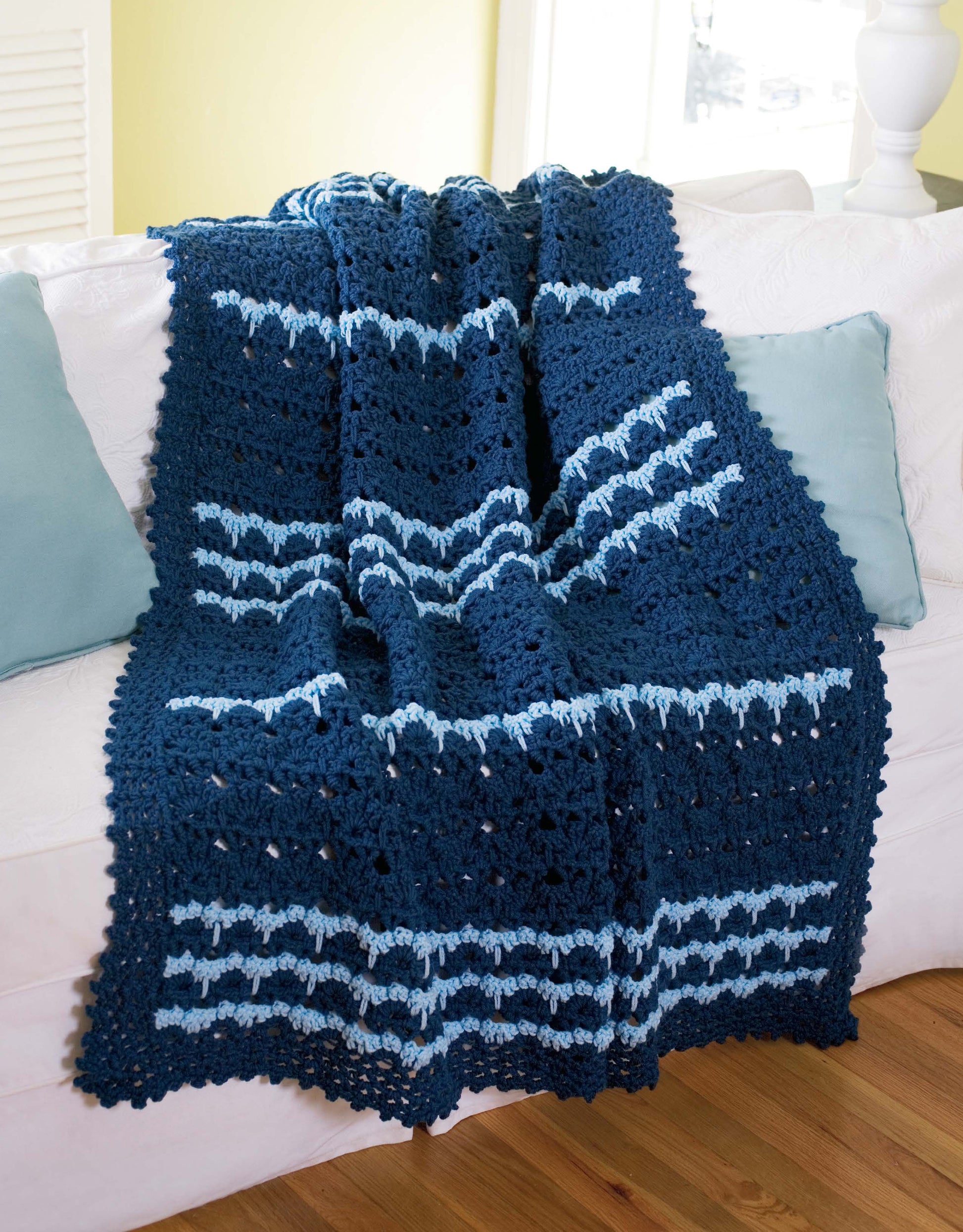The Contemporary Cottage Afghans Digital Download by Leisure Arts features a dark blue worsted weight crocheted blanket with light blue stripes draped over a white couch, accented by two light blue pillows and a white lamp in the background.