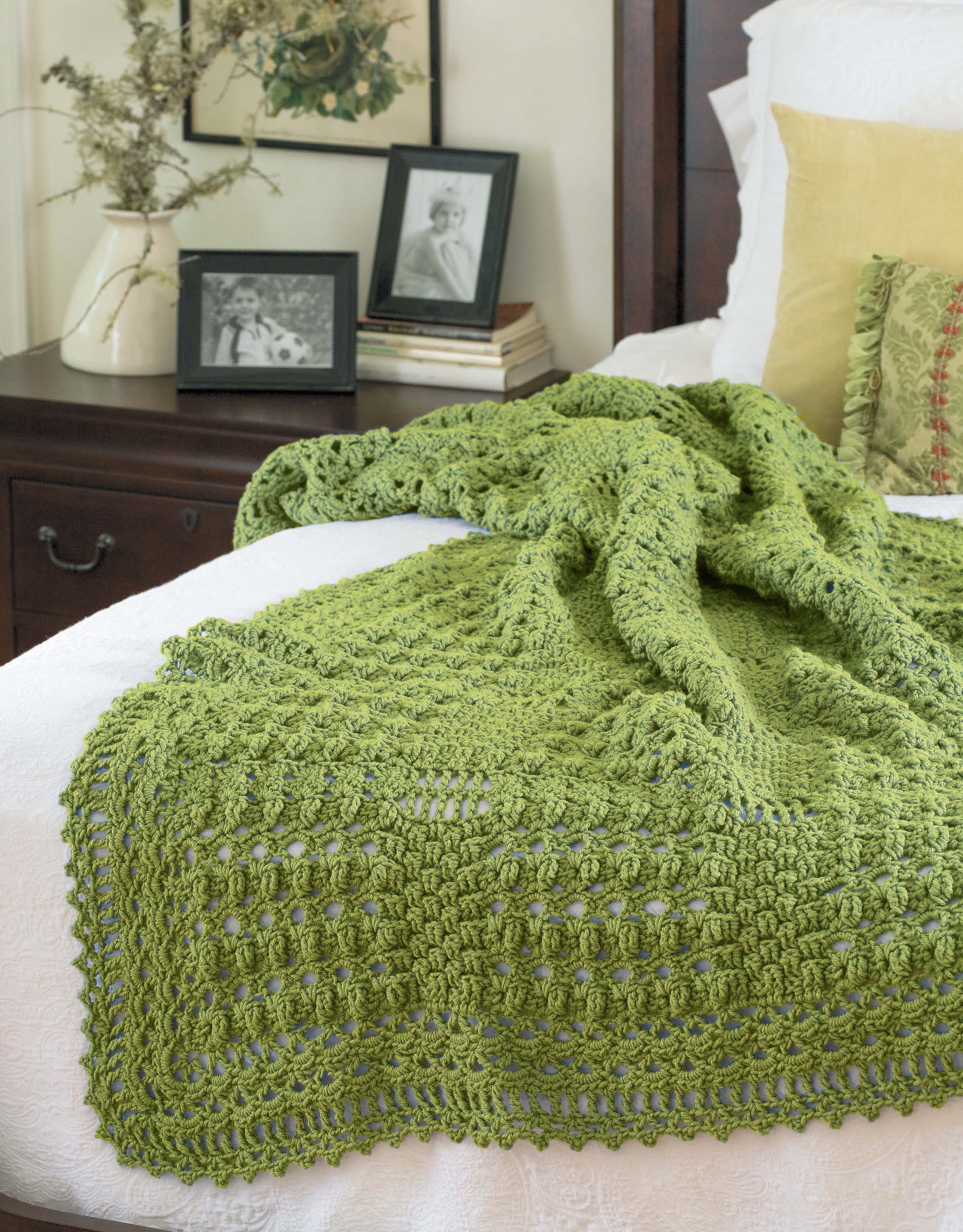 The Contemporary Cottage Afghans Digital Download by Leisure Arts features a textured green crocheted blanket made with worsted weight yarn, shown draped over a white bedspread beside a yellow pillow and decor on a wooden nightstand.