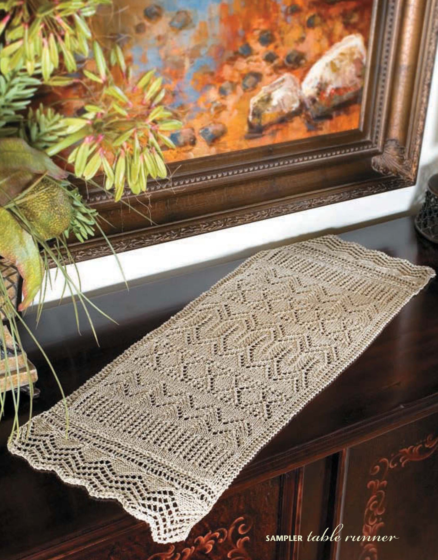 A hand-crocheted beige table runner with geometric patterns, inspired by "I Can't Believe I'm Lace Knitting" by Leisure Arts, lies on a dark wooden table beneath an earth-toned painting with green foliage.