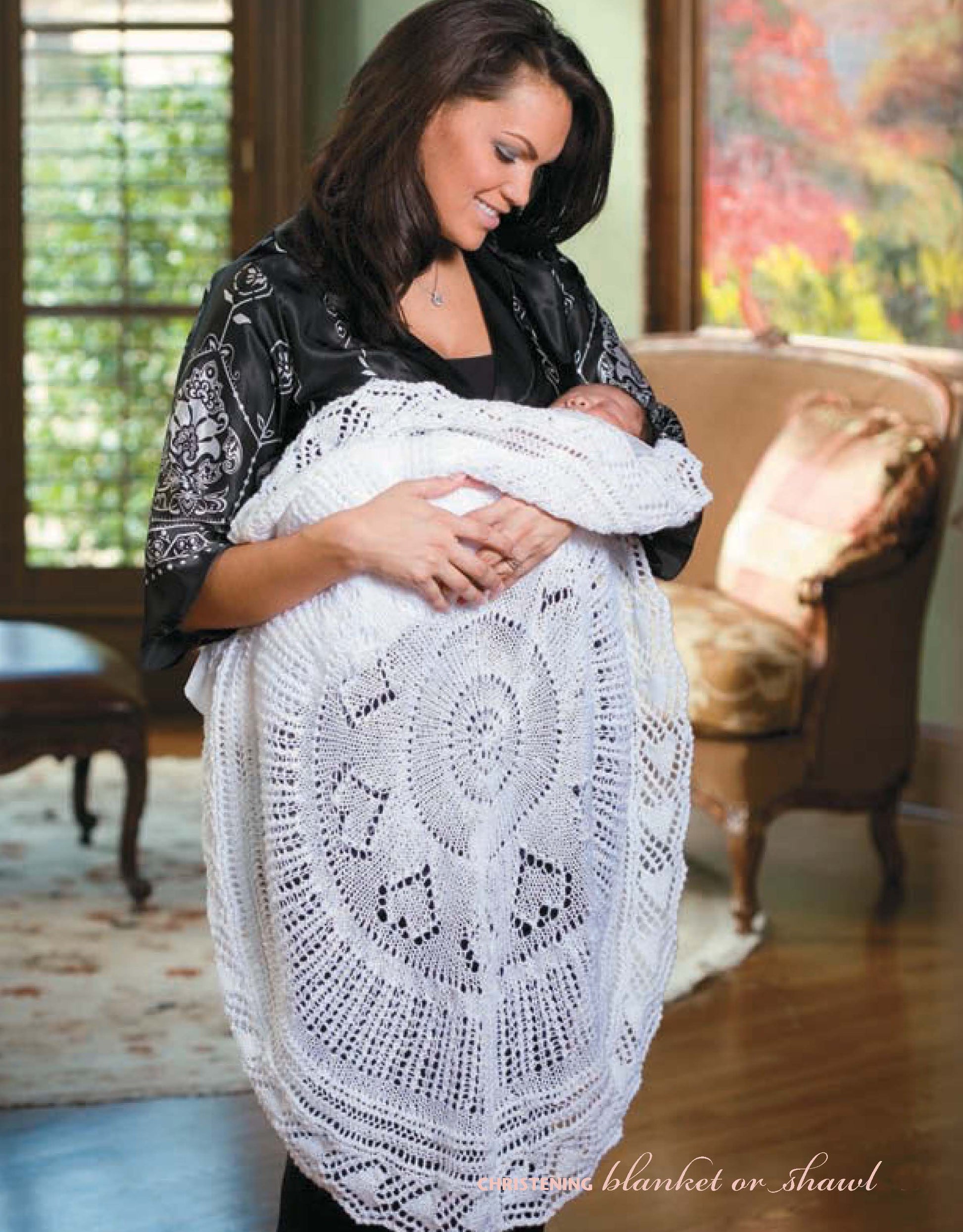 A woman smiles in a sunlit living room while holding a baby wrapped in a large white knitted blanket inspired by "I Can't Believe I'm Lace Knitting" from Leisure Arts.
