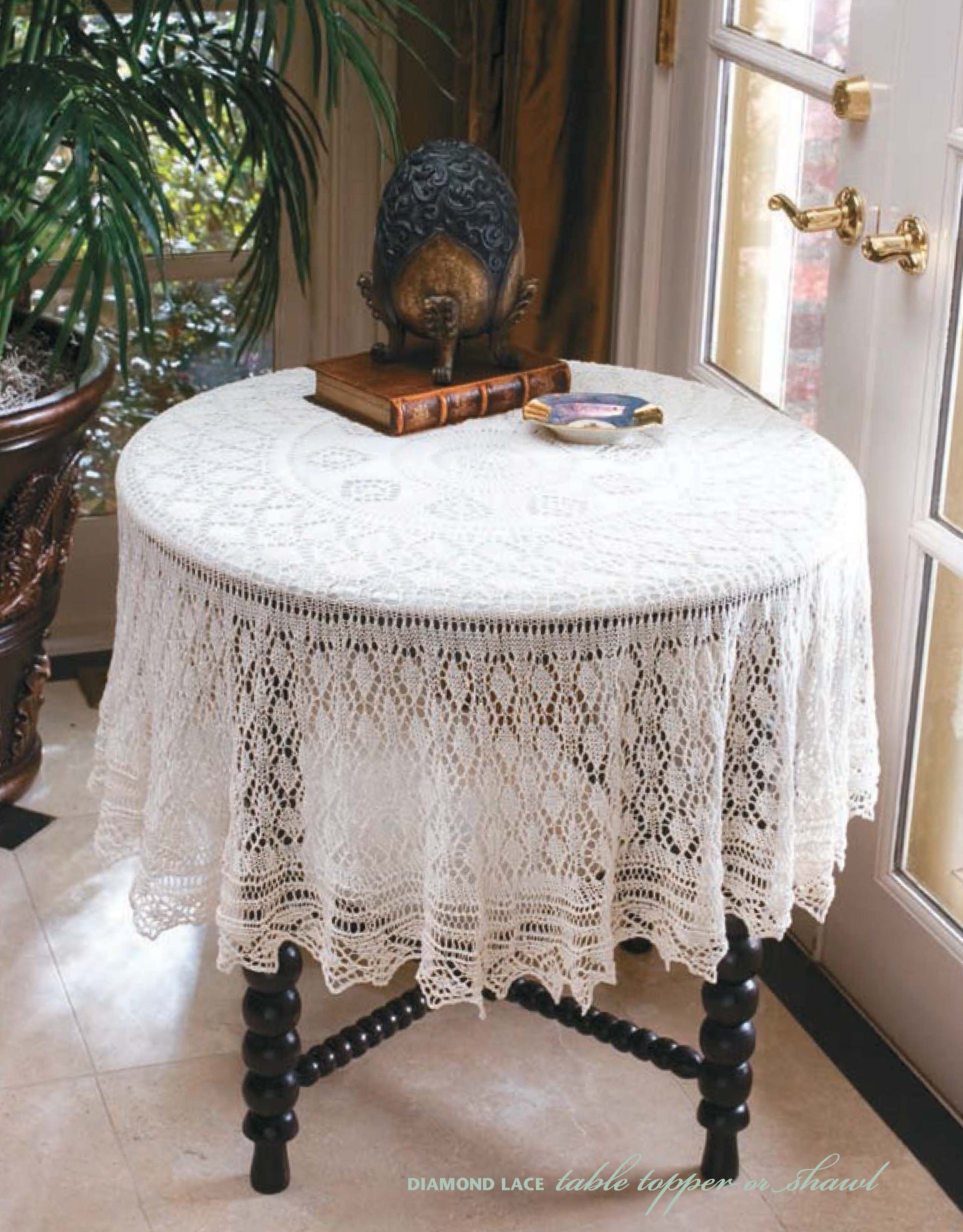 A round table with a white lace tablecloth inspired by Leisure Arts' "I Can't Believe I'm Lace Knitting" holds a decorative globe, books, and a dish. Sunlight streams in through nearby glass doors.