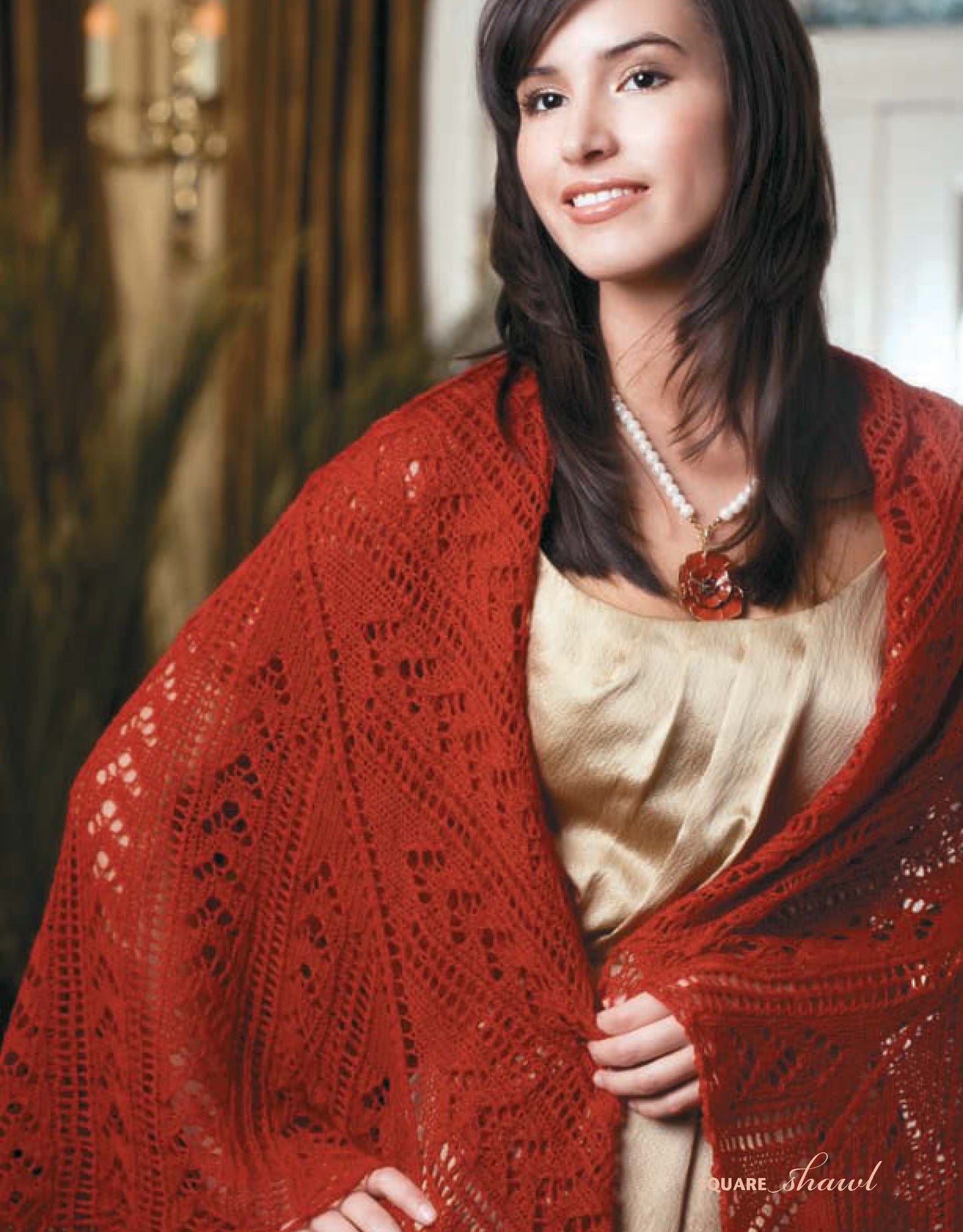 A woman with straight brown hair smiles in a light dress and a red lace shawl, featuring patterns from "I Can't Believe I'm Lace Knitting" by Leisure Arts. She accessorizes with a pearl necklace and a large red flower pendant.