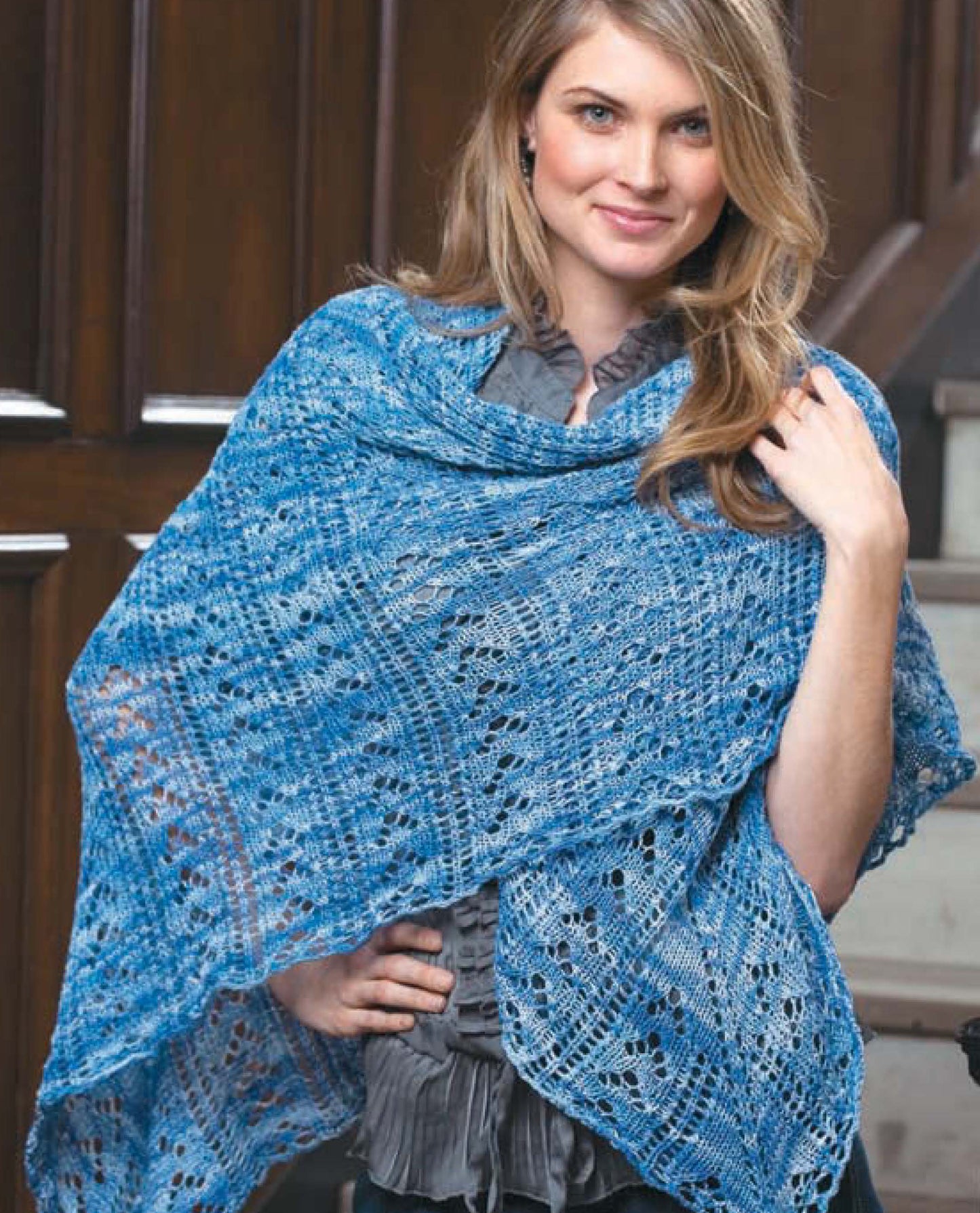A woman with long blonde hair smiles indoors, wearing a blue lace-knit shawl from Leisure Arts' "I Can't Believe I'm Lace Knitting" over a ruffled gray blouse. Wooden stairs and paneling can be seen in the background.