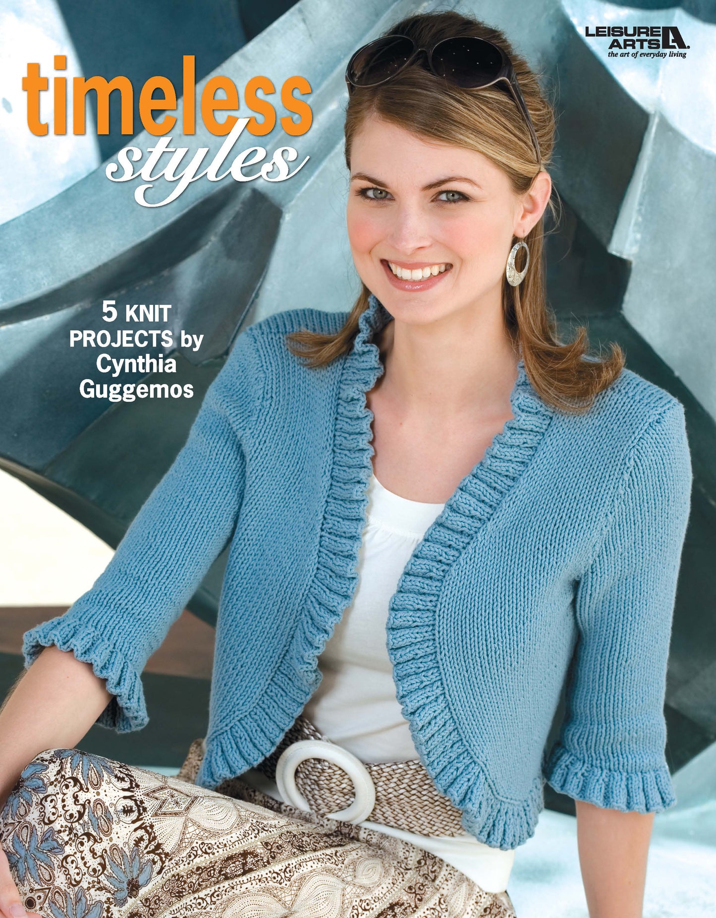 A smiling woman in a light blue knit cardigan and patterned skirt poses before an abstract background. Text reads “Timeless Styles—5 Knit Projects by Leisure Arts.”.