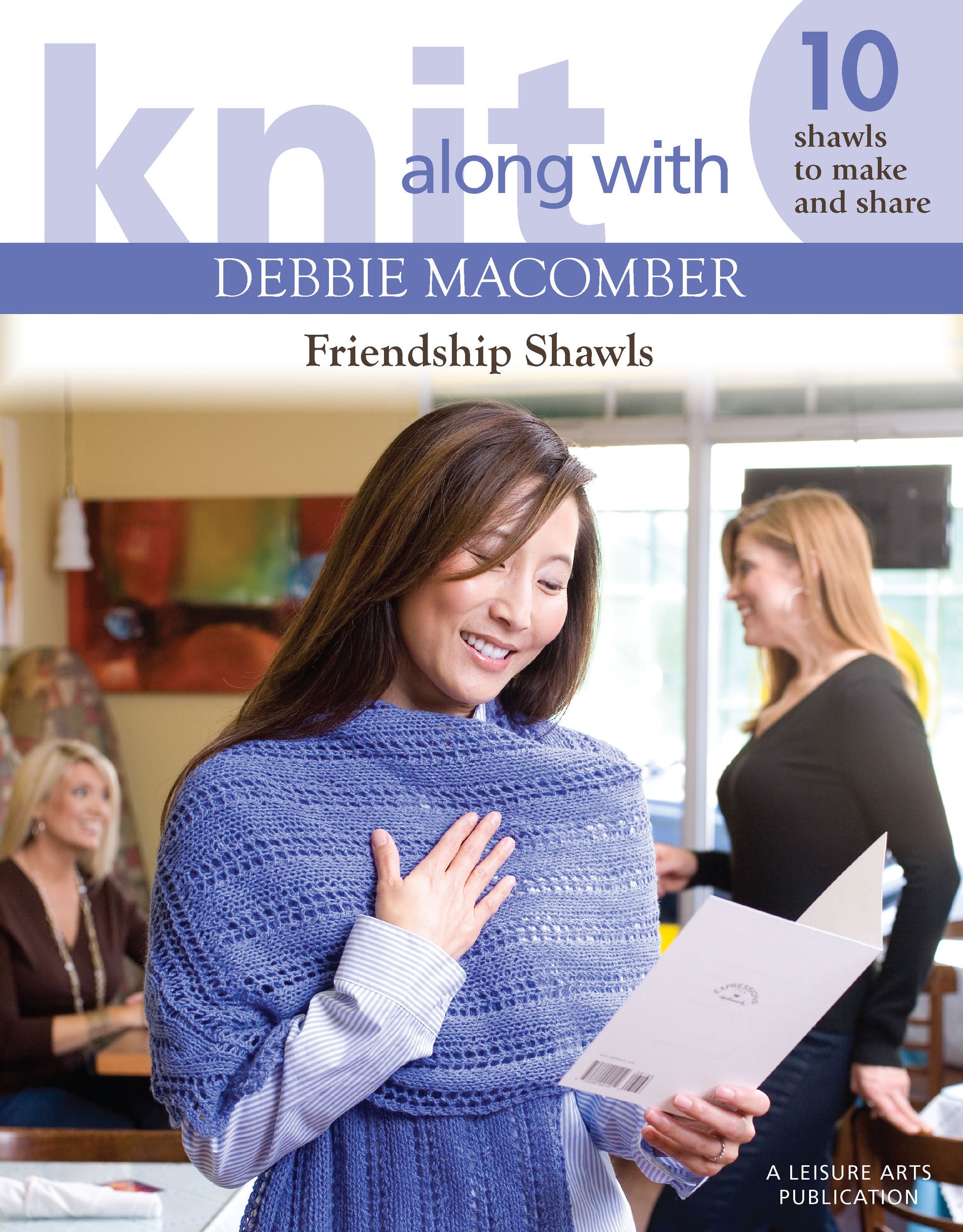 A smiling woman in a lavender shawl reads a card, while two women chat behind her. Cozy and bright, this Leisure Arts cover for Knit Along with Debbie Macomber—Friendship Shawls celebrates handmade gifts and friendship.