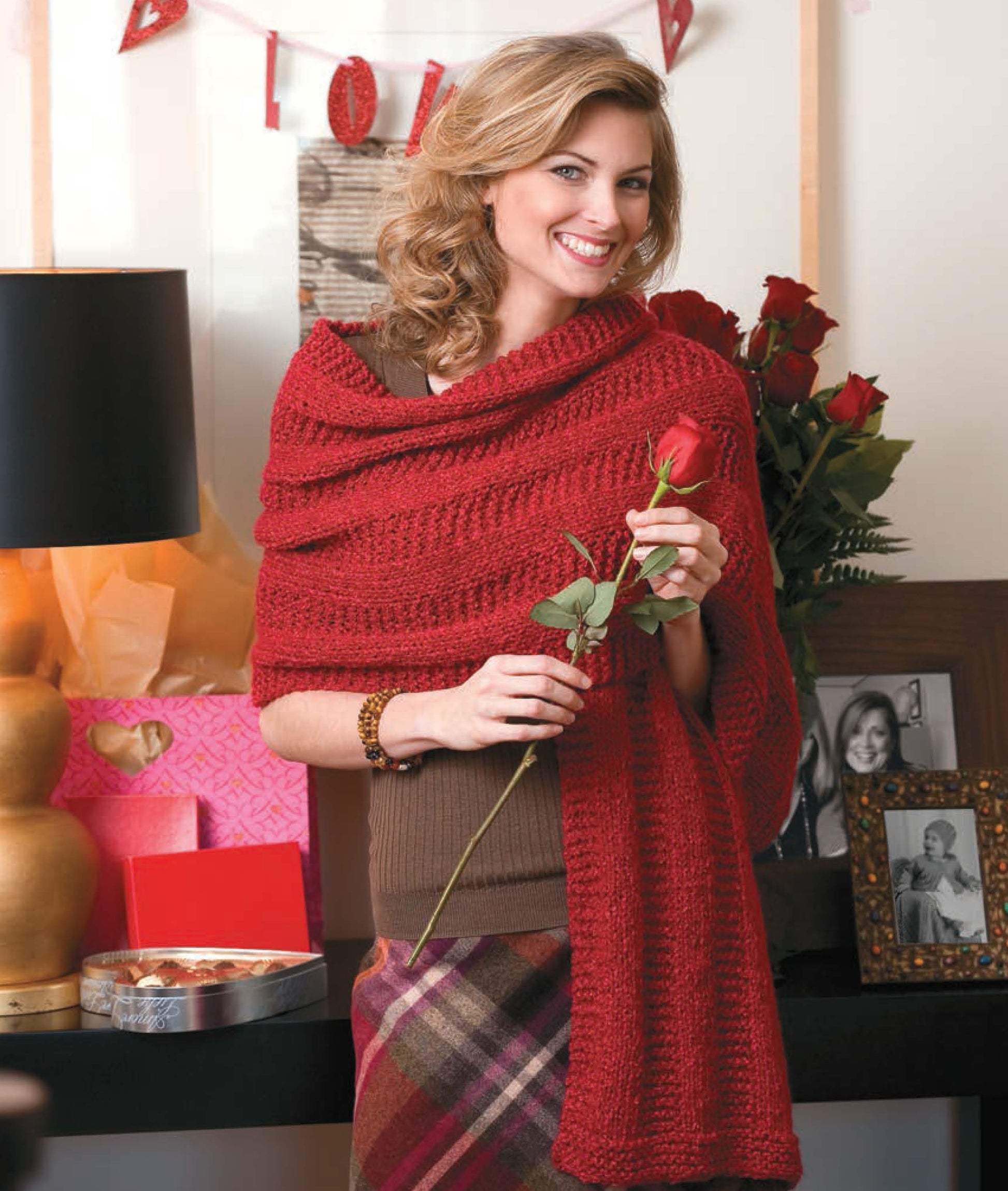 A smiling woman wears a red shawl made with "Knit Along with Debbie Macomber—Friendship Shawls" by Leisure Arts, holding a red rose. Red roses, a LOVE banner, handmade gifts, family photos, and heart chocolates create a festive, romantic scene.