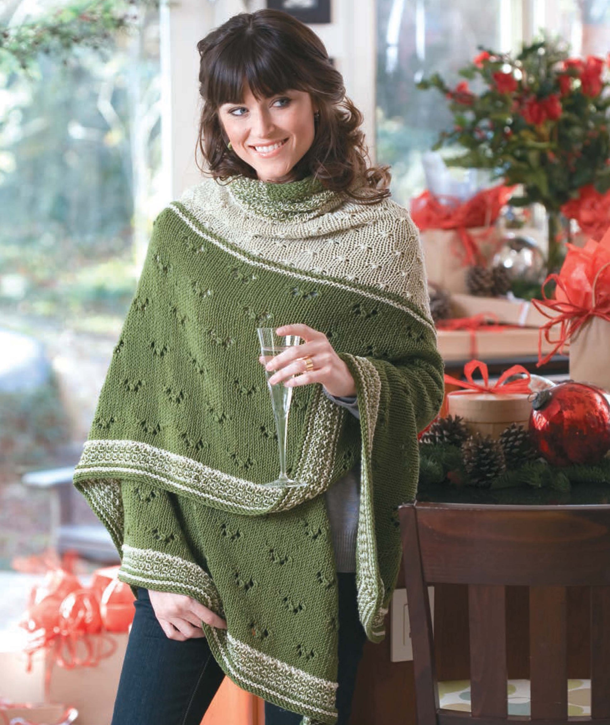 A woman smiles indoors, holding a glass and wearing a shawl made from the Leisure Arts "Knit Along with Debbie Macomber—Friendship Shawls" kit. Behind her are wrapped handmade gifts, festive decorations, and red bows for a holiday feel.