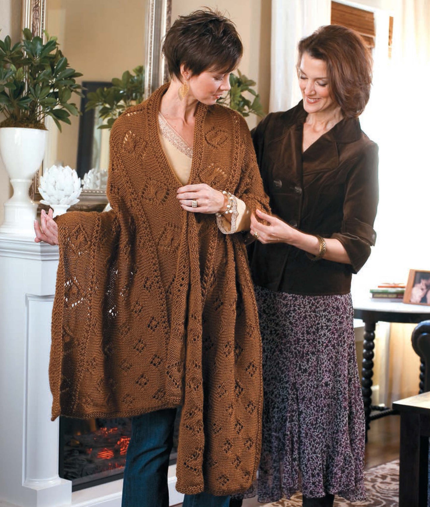 In a cozy living room, one woman models the Leisure Arts "Knit Along with Debbie Macomber—Friendship Shawls," displaying its brown diamond-knit pattern as her friend admires and gently holds the edge. Both smile, reflecting the joy of handmade gifts.