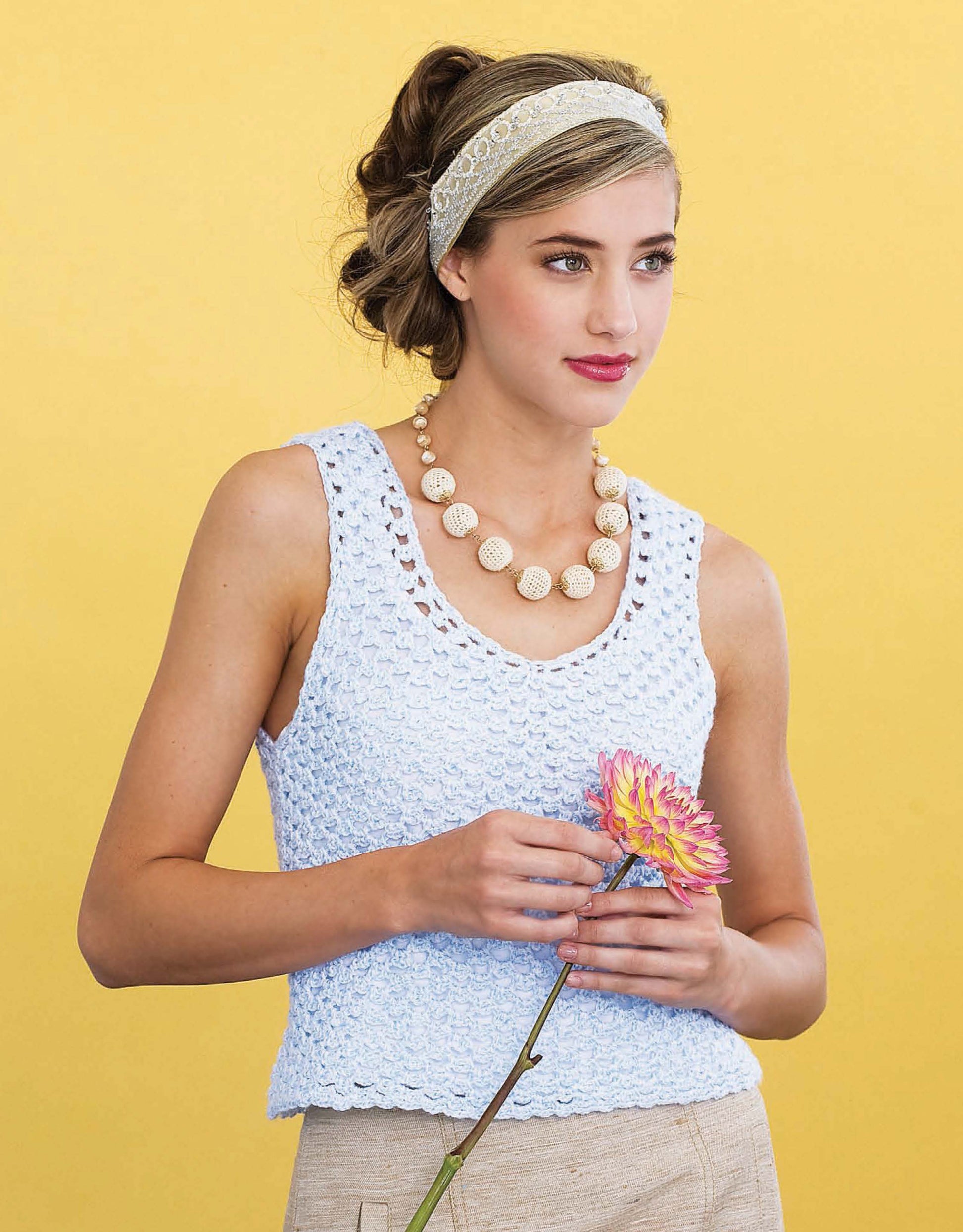 A woman models the Crochet Easygoing Fashions top by Leisure Arts, styled with beige pants, a chunky beaded necklace, and a lace headband as she holds a pink and yellow flower against a yellow background.