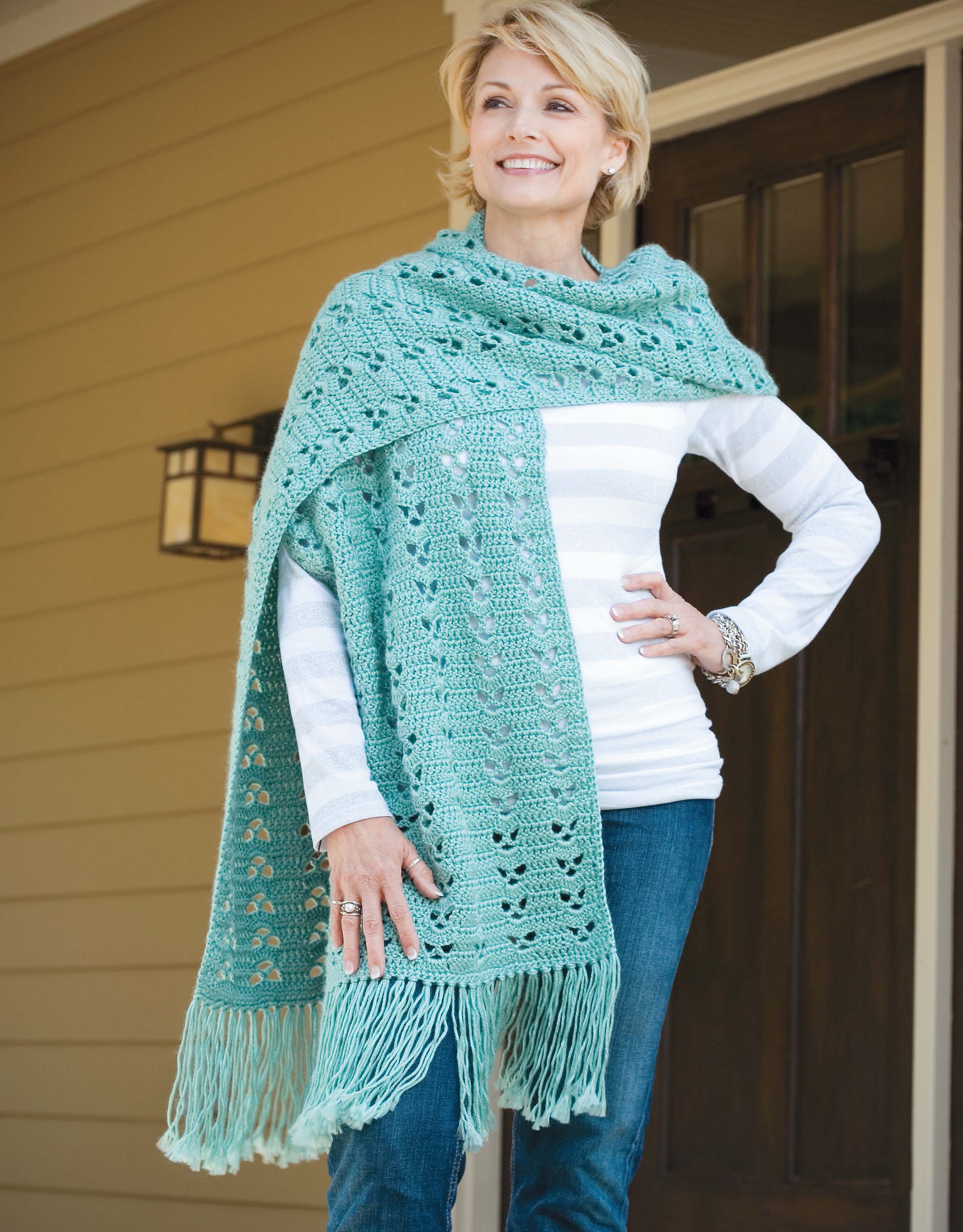 A woman on a porch smiles, wearing a light green lacy shawl with fringe over a striped shirt and jeans. Made with Leisure Arts' "Learn a Stitch Create a Scarf Digital Download," this crochet pattern adds stylish flair to any outfit.
