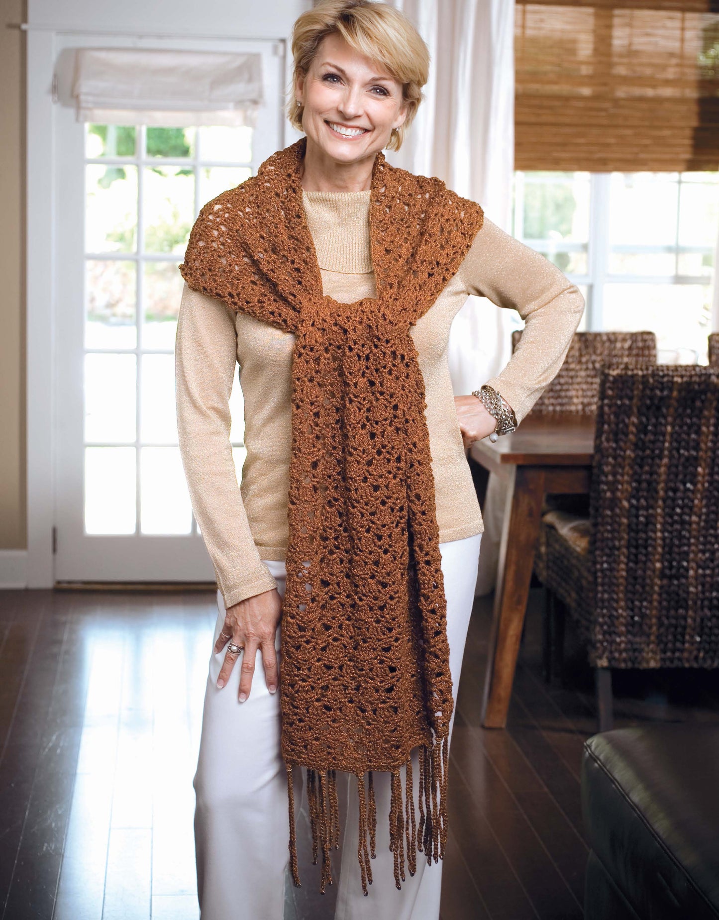 A woman smiles indoors, modeling a beige turtleneck, white pants, and a brown crocheted scarf with fringe made using the "Learn a Stitch Create a Scarf Digital Download" by Leisure Arts in a bright room with wood floors and modern décor.