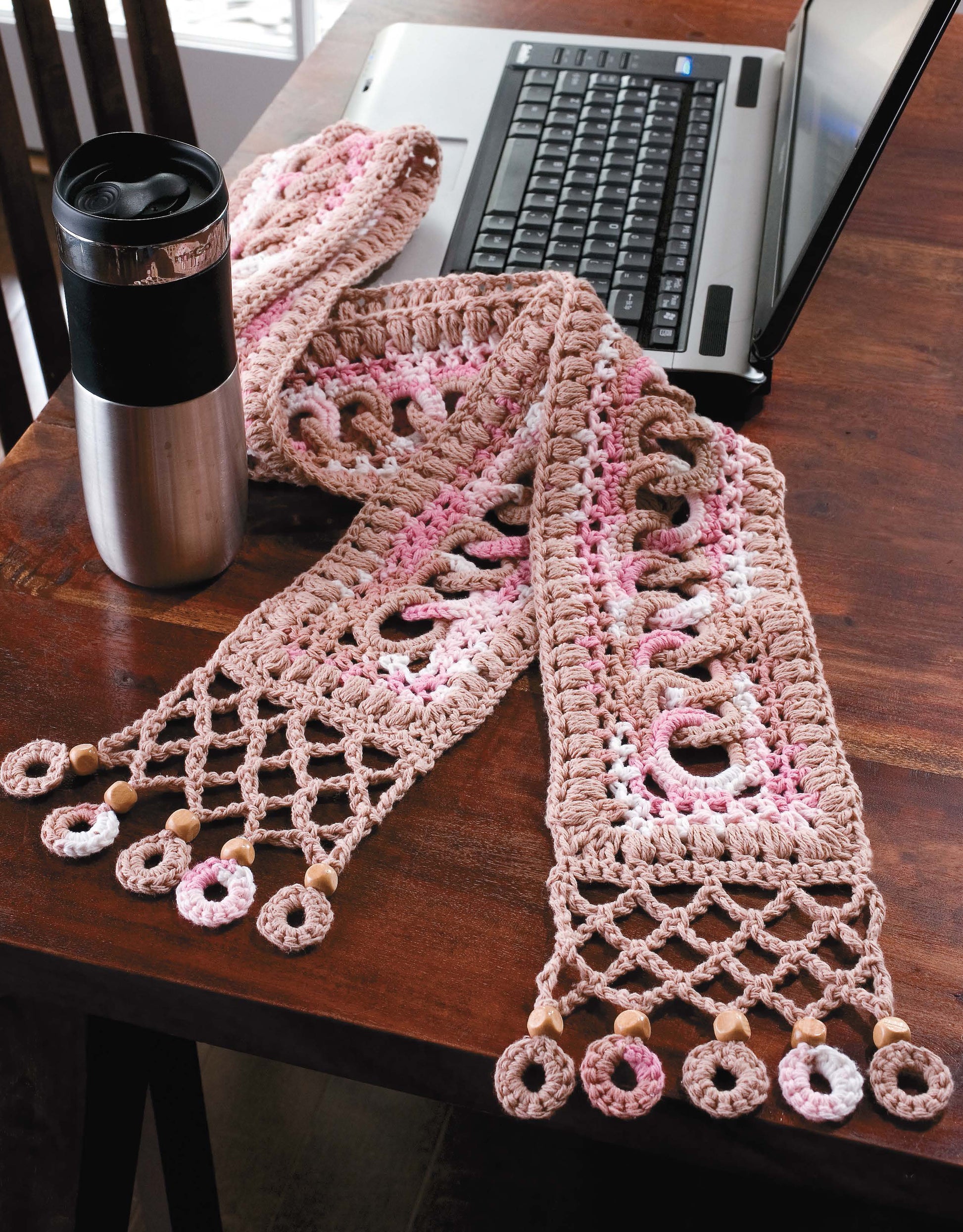 The Learn a Stitch Create a Scarf Digital Download by Leisure Arts features pink and beige crochet with wooden bead accents, inspired by afghan patterns—shown draped near a travel mug and open laptop on a wooden table.