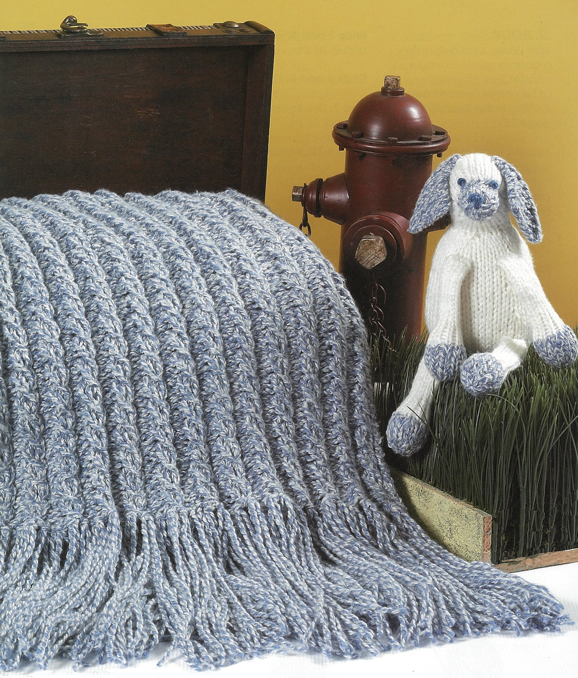 A Leisure Arts "A Collection of Characters" light blue and gray fringed knitted blanket is draped over a wooden chest with a matching knit white dog toy, next to a red fire hydrant, all set against a yellow background.