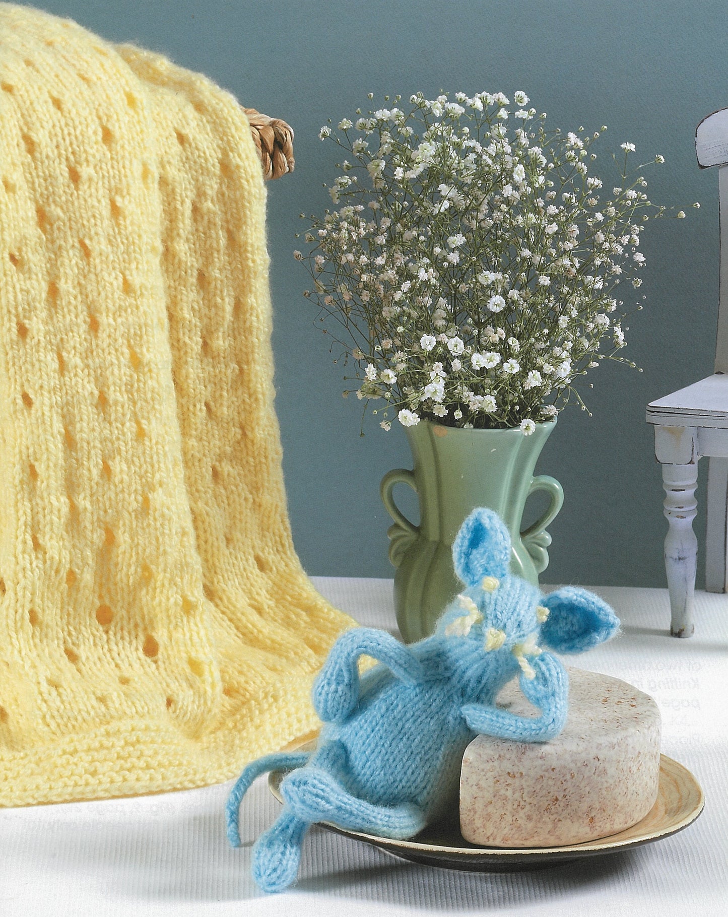 A soft yellow knit blanket from Leisure Arts' "A Collection of Characters" set hangs beside a green vase with white flowers. A blue knitted mouse sits on soap by a white chair, all set against a blue background.