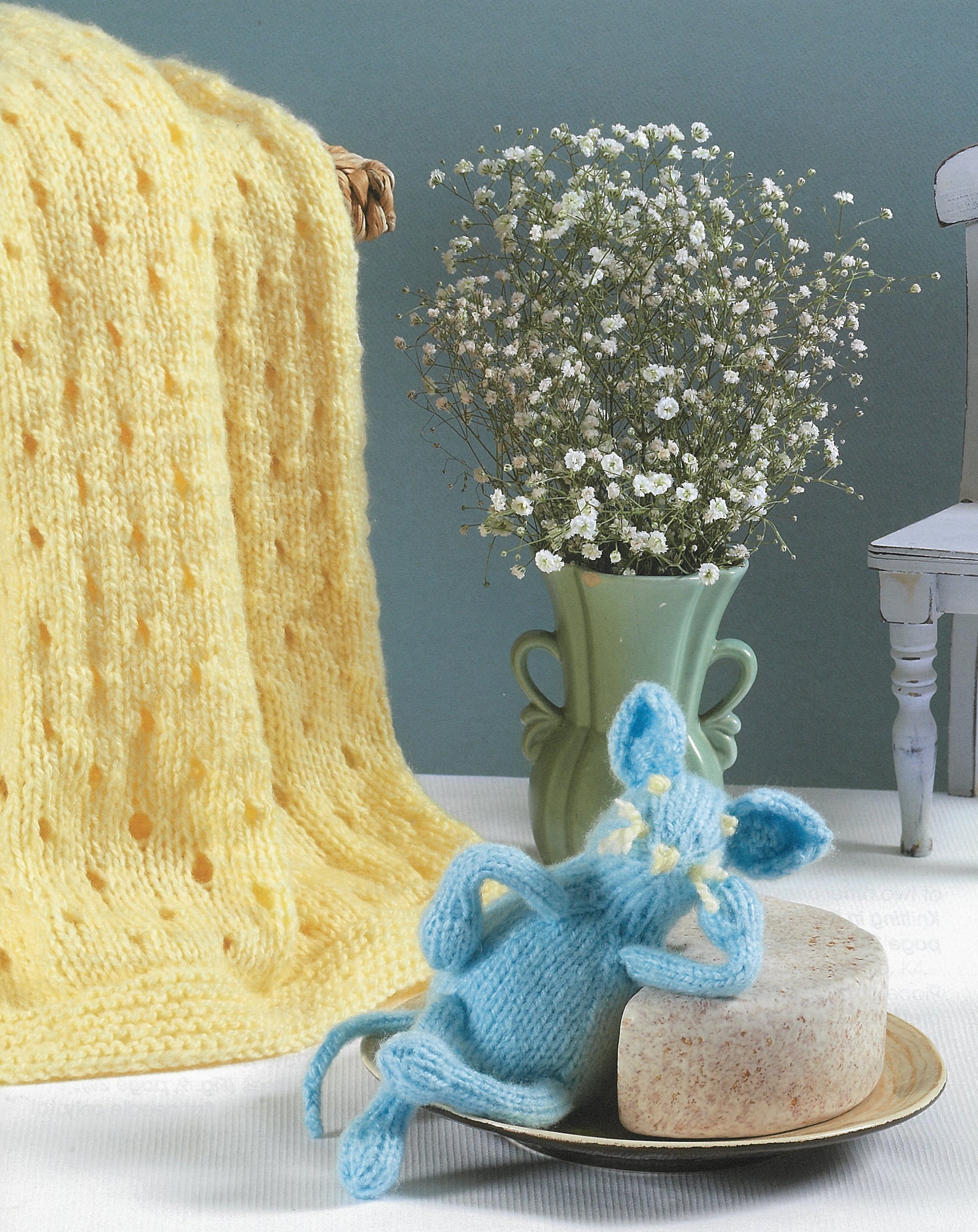 A soft yellow knit blanket from Leisure Arts' "A Collection of Characters" set hangs beside a green vase with white flowers. A blue knitted mouse sits on soap by a white chair, all set against a blue background.