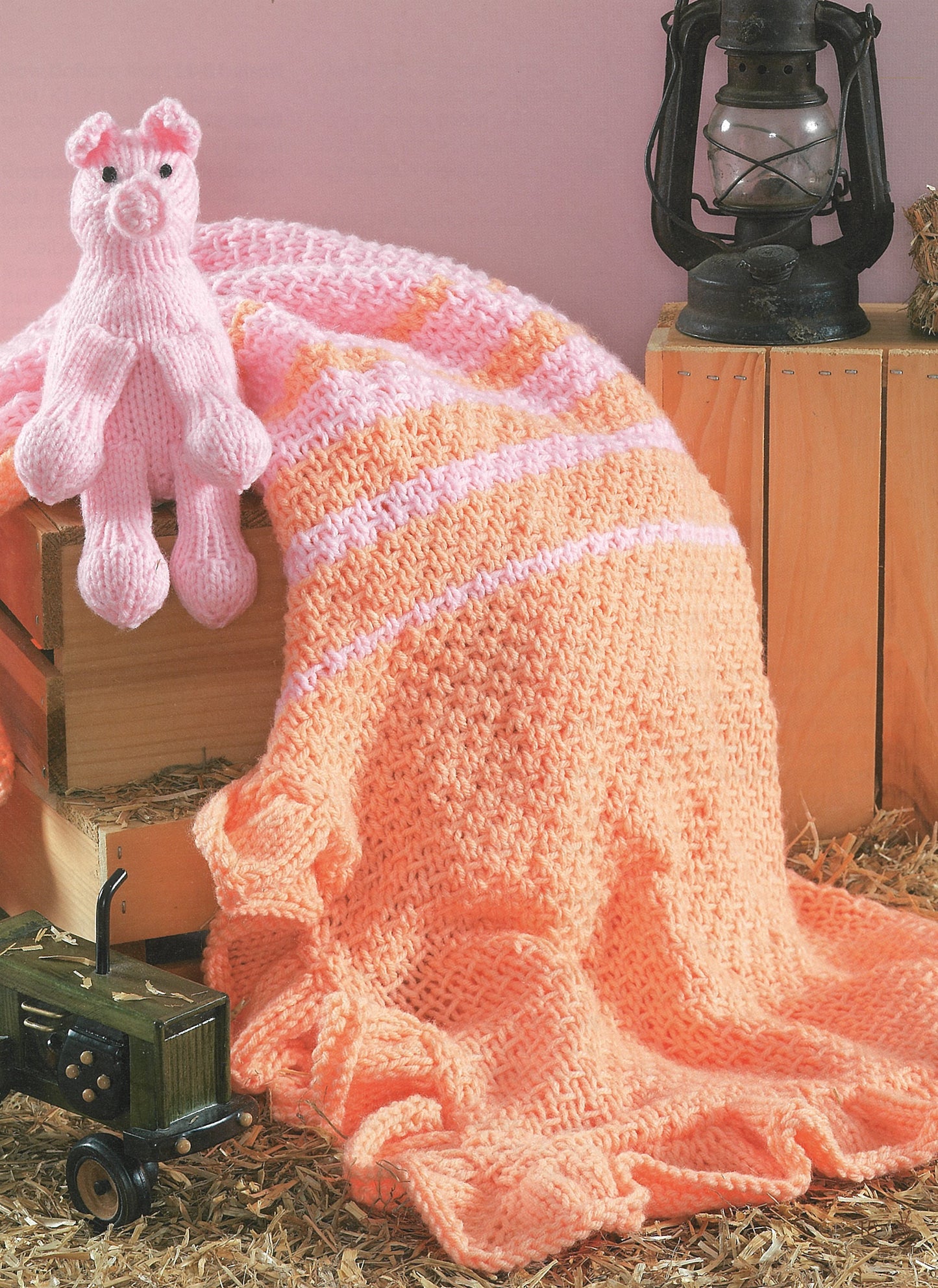 A Collection of Characters by Leisure Arts features a soft peach and pink knit blanket draped on a wooden crate with a pink pig toy, set beside a vintage lantern and toy truck on straw against a pink backdrop.